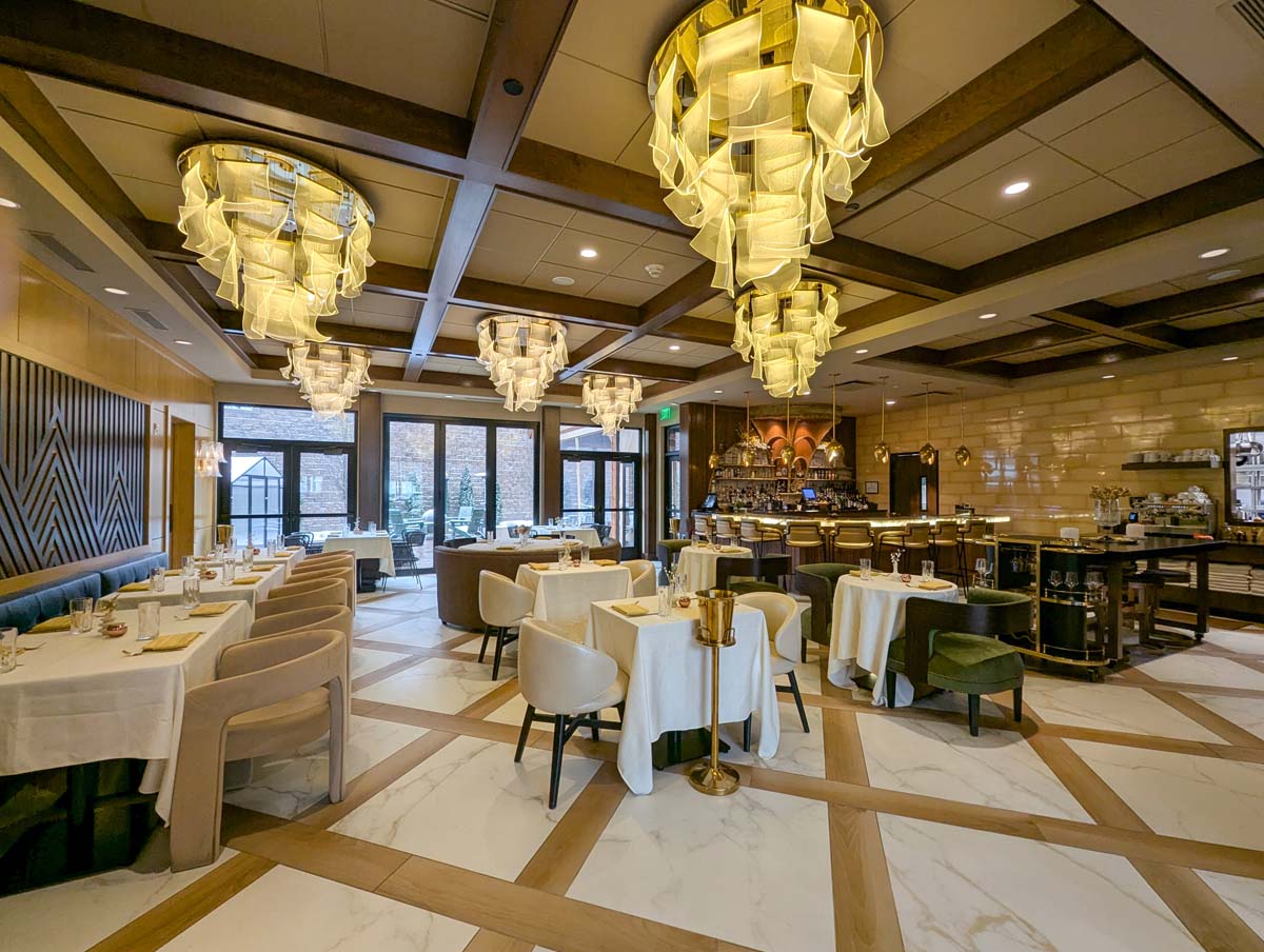 Sophisticated restaurant interior featuring dramatic cascading gold chandeliers, marble and wood inlay flooring, white tablecloths, modern cream and green upholstered chairs, full-service bar, and floor-to-ceiling windows overlooking the property