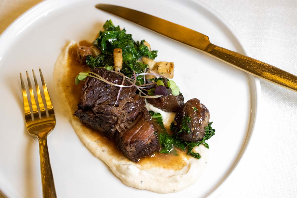 Elegantly plated braised short rib on white ceramic plate with celery root puree, sautéed kale, roasted pearl onions, microgreens, and rich demi-glace sauce, served with gold-plated cutlery