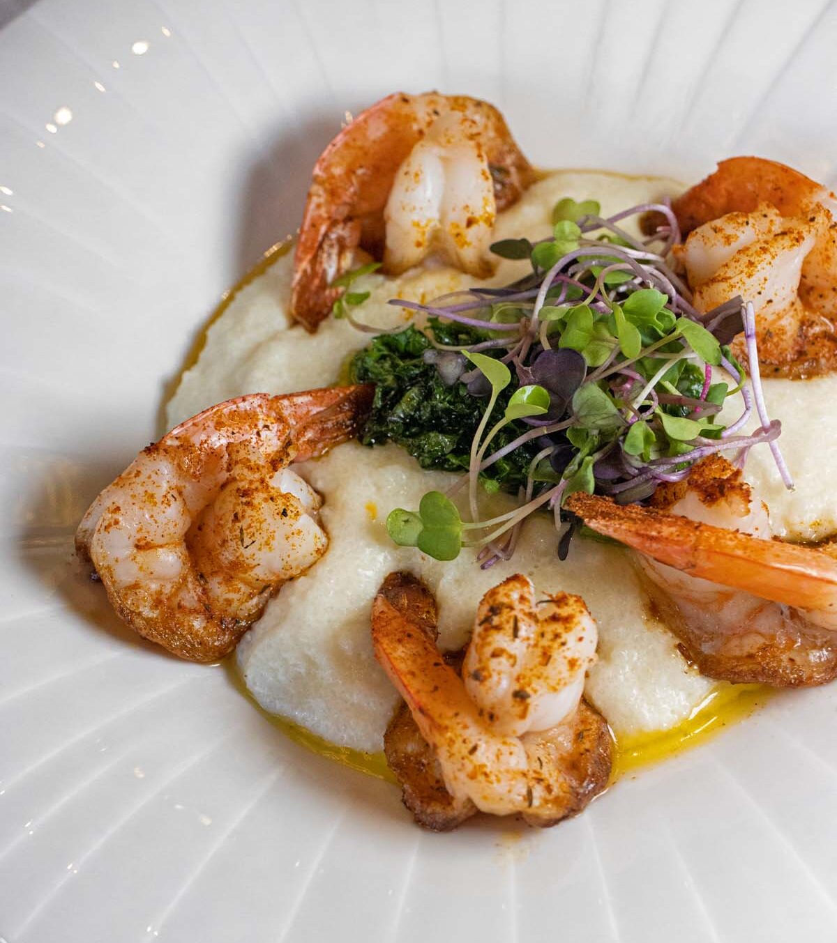 Artfully plated Shrimp and Grits featuring perfectly seared jumbo shrimp, creamy grits, sautéed kale, and colorful microgreens with purple and green accents, served on a white ribbed plate with olive oil drizzle