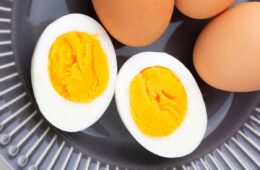 Two halved hard-boiled eggs showing bright yellow, creamy yolks and firm white albumen, placed on a gray ridged surface with uncooked brown eggs in the background