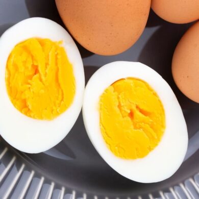 Two halved hard-boiled eggs showing bright yellow, creamy yolks and firm white albumen, placed on a gray ridged surface with uncooked brown eggs in the background
