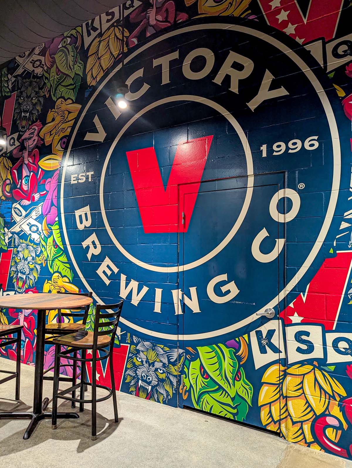 Large brewery logo mural painted on a navy blue wall featuring Victory's signature 'V' in red, surrounded by vibrant street art-style illustrations of hops, wolves, and geometric designs. A high-top bar table and stools are positioned in front of the mural.