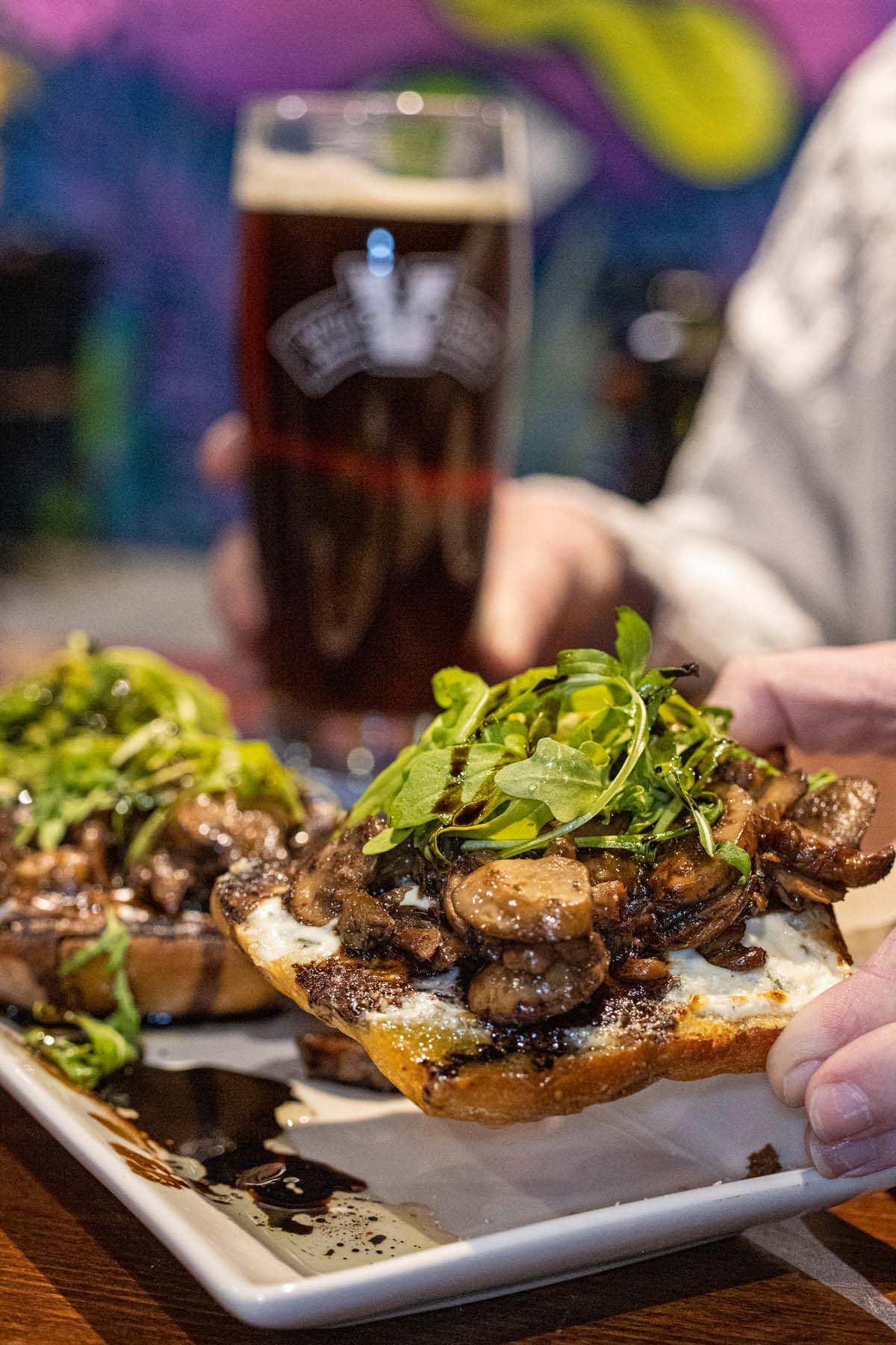 Artisanal open-faced sandwich featuring sautéed mushrooms topped with fresh arugula on toasted bread, served on a white plate. A Victory Brewing Company beer glass with a dark brew is visible in the background against colorful wall art