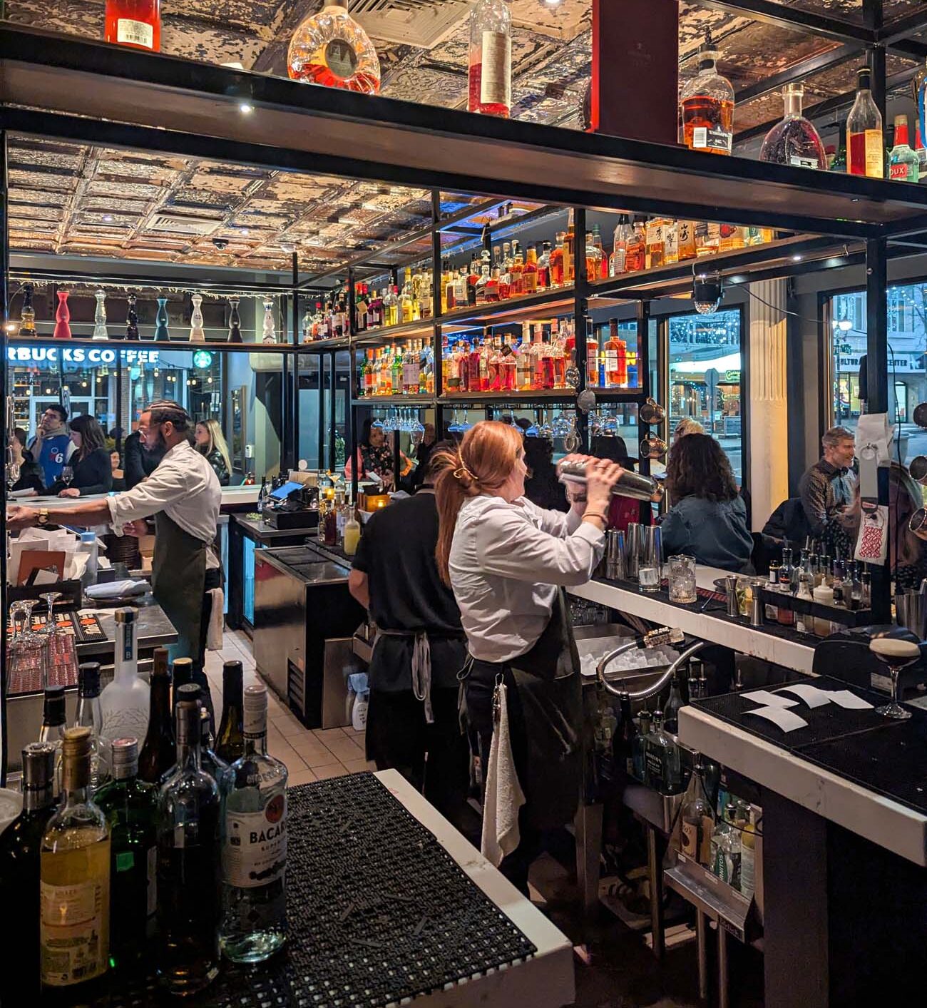 A busy, well-stocked bar at Bardea Food & Drink featuring an ornate tin ceiling with distressed gold patina. Bartenders in white shirts and black aprons mix drinks while patrons fill the space. Multiple shelves display colorful bottles of spirits, with special attention to amber and red-colored liquors. The warm lighting and industrial-chic design create an inviting atmosphere.