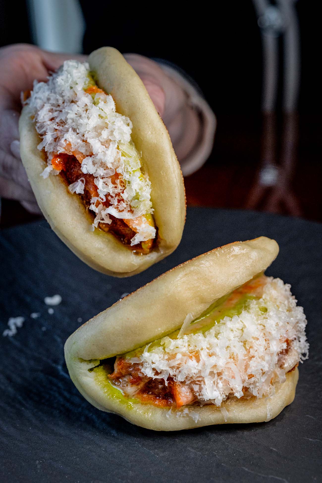 wo pillowy white bao buns on a black slate surface. Each bun is filled with crispy chicken parm topped with shredded mozzarella cheese and vodka sauce. The filling peeks out from the folded buns, showcasing the Italian-Asian fusion concept that Bardea is known for.