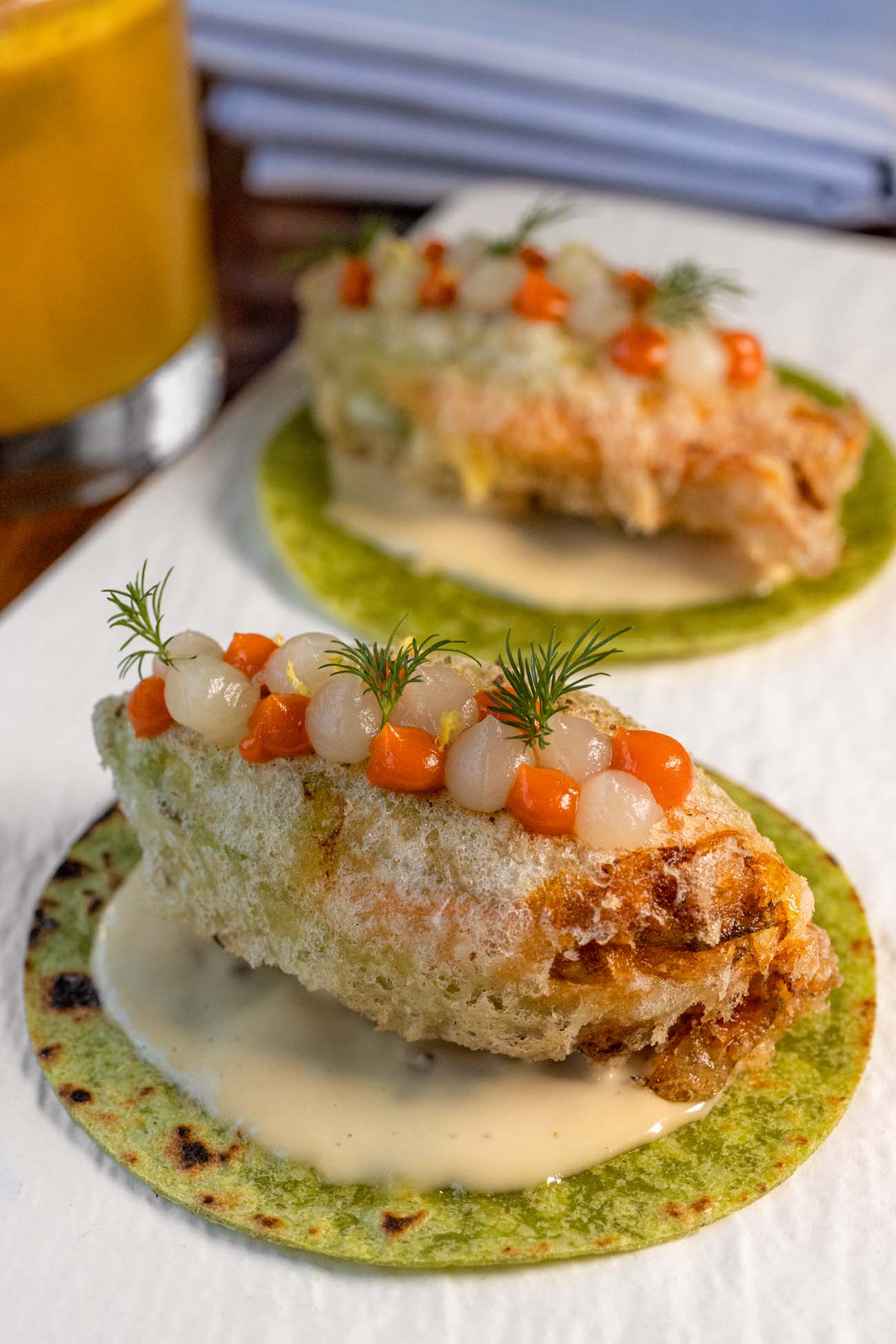 Two artfully plated crab tacos on a white plate. Each features a vibrant green tortilla base topped with tempura-fried squash blossoms stuffed with crab filling. The tacos are garnished with fresh dill fronds and dots of creamy sauce, with an orange-yellow Working Girl cocktail visible in the background.