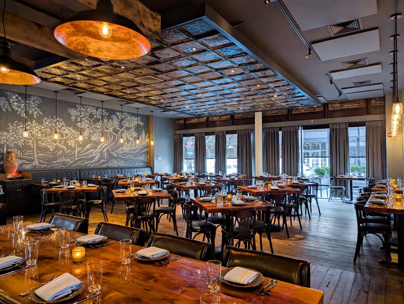 A spacious dining room with warm lighting and rustic elegance. Features wooden tables with black bentwood chairs, textured tin ceiling tiles, copper pendant lighting, and a striking wall mural depicting trees. Large windows with gray curtains line one wall, while place settings with glasses and napkins await diners. The atmosphere combines industrial and warm design elements.