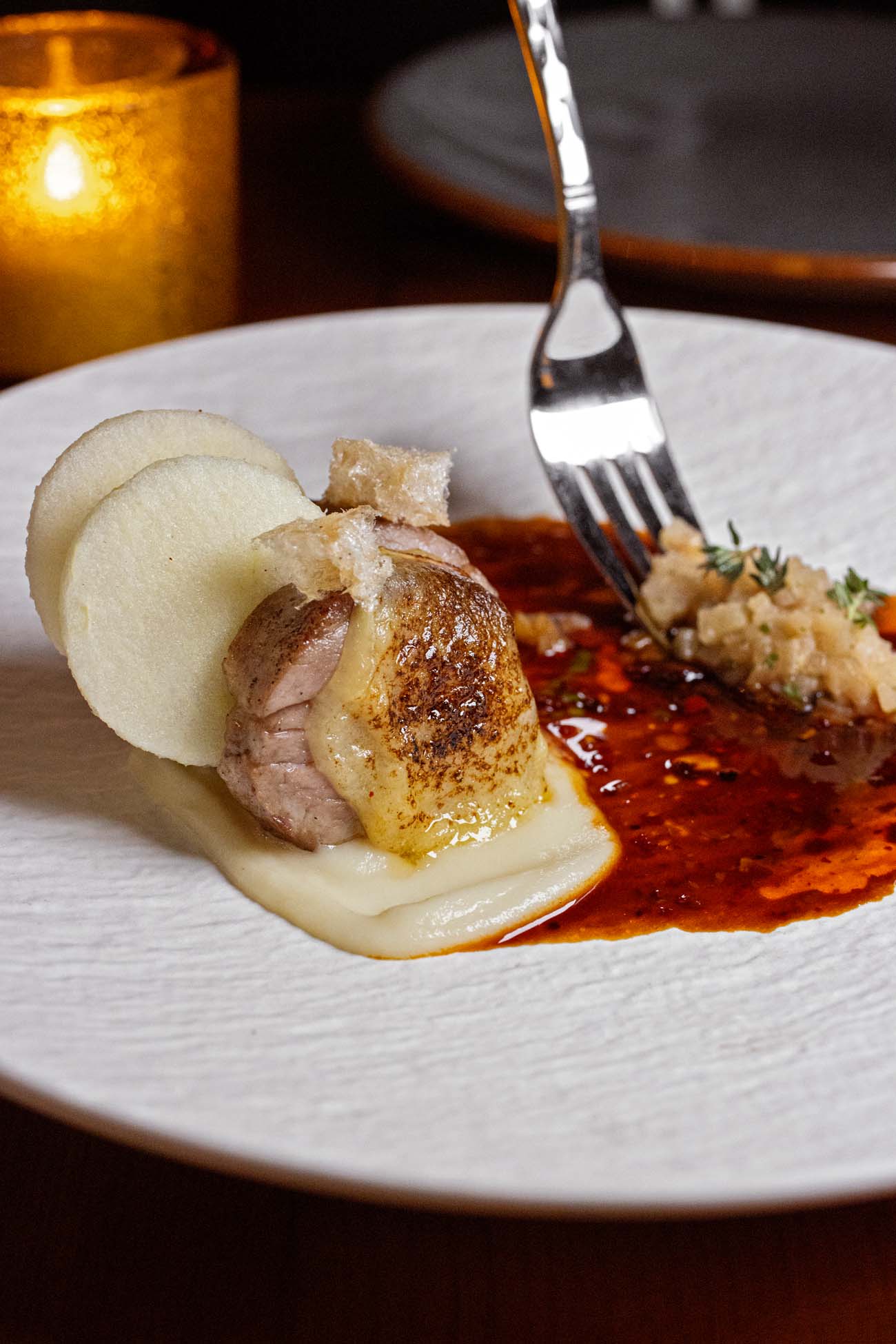 A beautifully plated Iberico pork medallion on a textured white plate. The pork is seared with a golden crust and sits atop a creamy sauce. Accompanied by thin slices of green apple, a rich truffle-chili au jus, and a small portion of what appears to be compote. A silver fork rests on the plate, poised to take a bite.