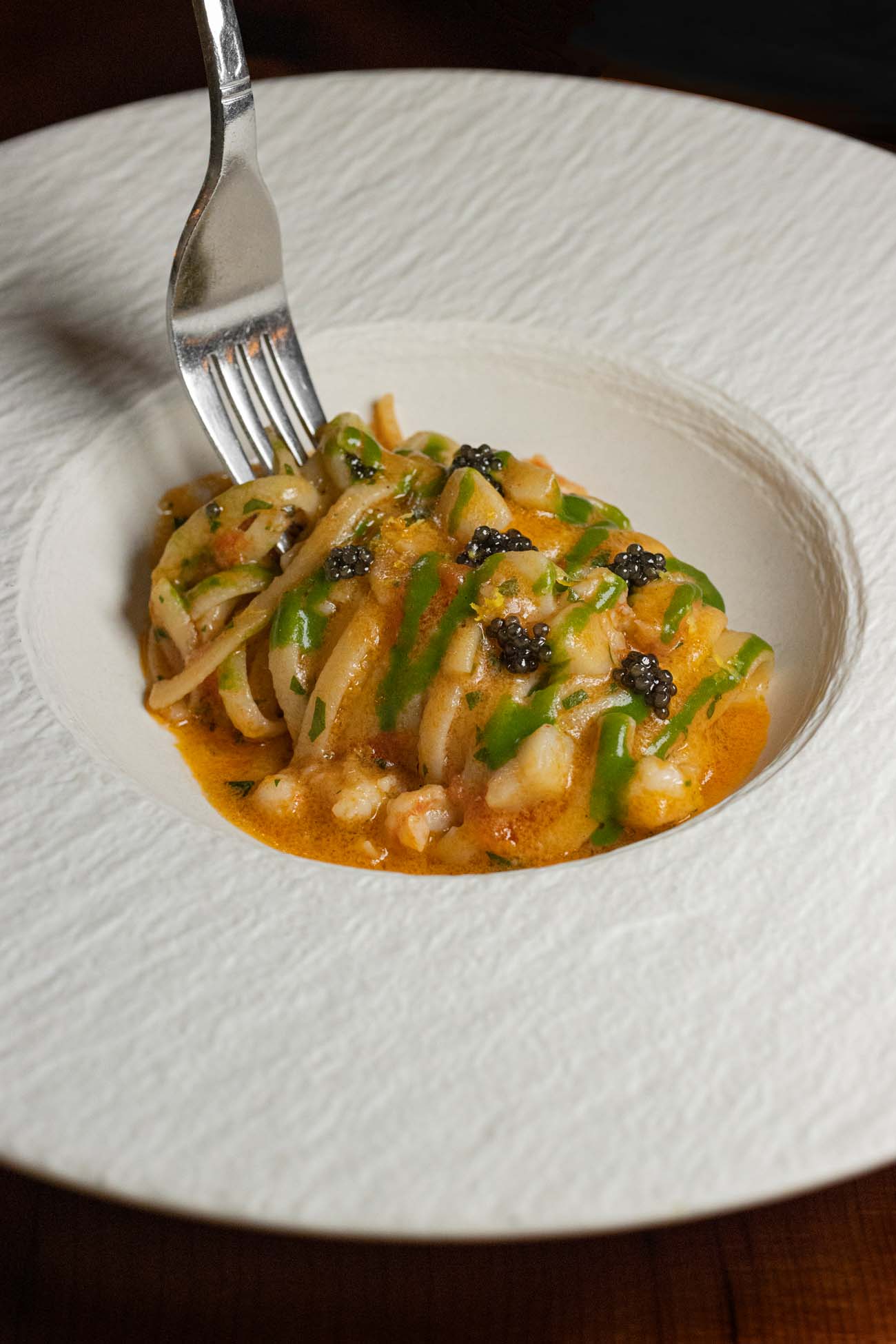 An elegantly plated pasta dish served in a textured white bowl containing handmade linguine with shrimp in a rich orange-hued sauce, garnished with small black caviar pearls and green herb drizzles. A silver fork rests at the edge of the bowl, displaying the luxurious pasta dish mentioned in the article as one of Bardea's decadent specialties.