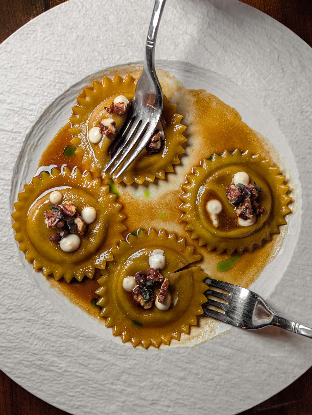 Four golden-colored, gear-shaped ravioli arranged on a white textured plate in amber broth. Each raviolo is topped with dots of white cheese cream and crunchy pecan pieces. Two silver cocktail forks rest on the plate, emphasizing the sharing nature of the dish. The pasta's unique scalloped edges create a sophisticated presentation.