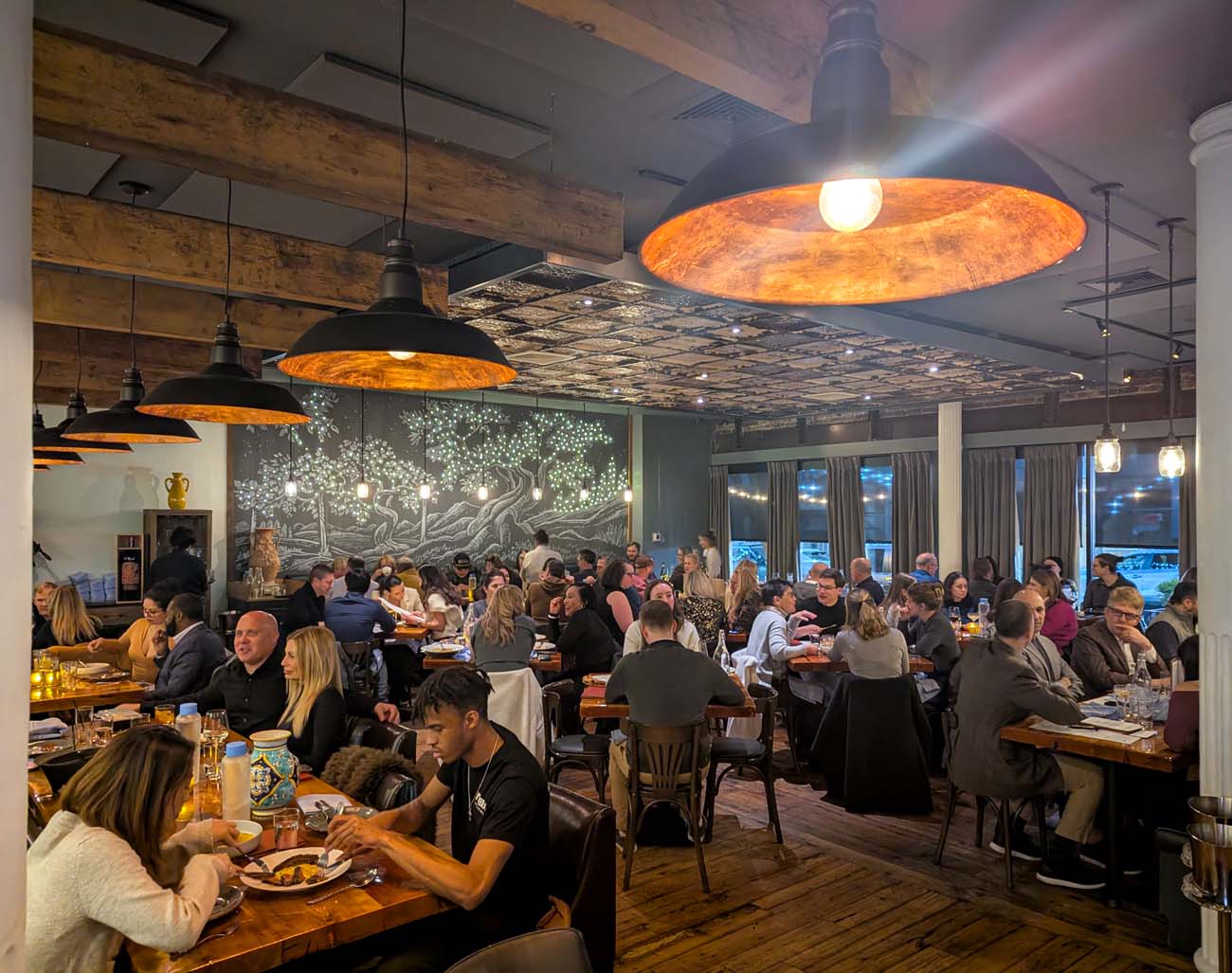 A vibrant, packed dining room at Bardea Food & Drink during dinner service. The space features rustic wooden tables filled with diners enjoying meals, industrial-chic copper pendant lights with warm orange interiors, exposed wooden beams, and a distinctive pressed tin ceiling. A striking wall mural of illuminated trees creates an artistic focal point against the gray wall. Large windows with gray curtains line one side, while the room buzzes with conversation as servers navigate between tables. The warm lighting and full tables convey the restaurant's popularity.