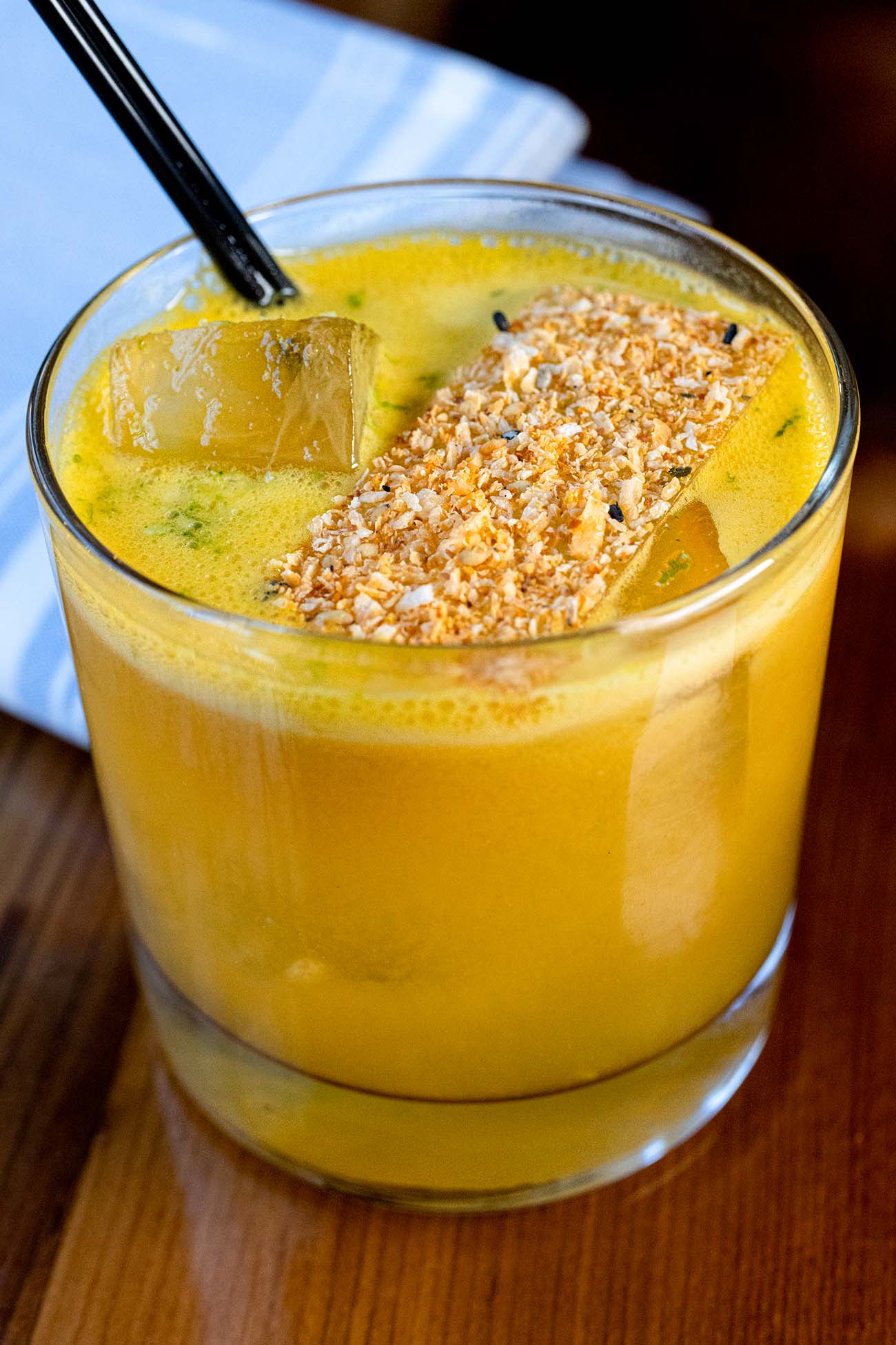 A close-up of Bardea's Working Girl cocktail in a rocks glass on a wooden surface. The drink has a vibrant golden-yellow color from its blend of tequila, passion fruit, and citrus. A large square ice cube sits on one side, while a rectangular isomalt garnish topped with toasted coconut and togarashi powder stretches across the top. A black straw enters the drink from the left, and hints of green herbs are visible in the liquid. The presentation showcases the bar's attention to detail and creative mixology.