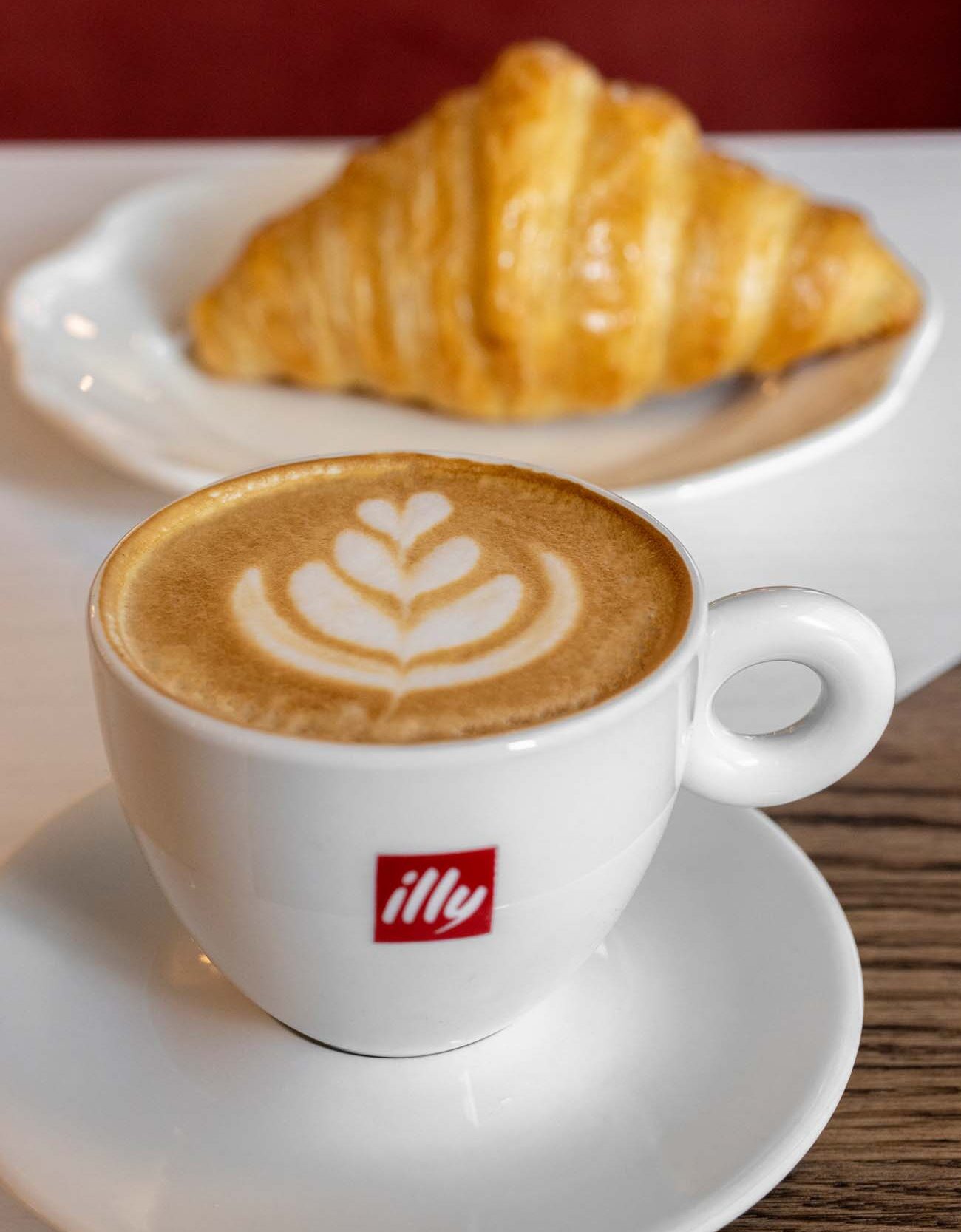 Close-up of a white Illy-branded cappuccino cup containing coffee with a decorative leaf pattern in the foam. In the background, slightly blurred, is a golden-brown croissant on a white plate.