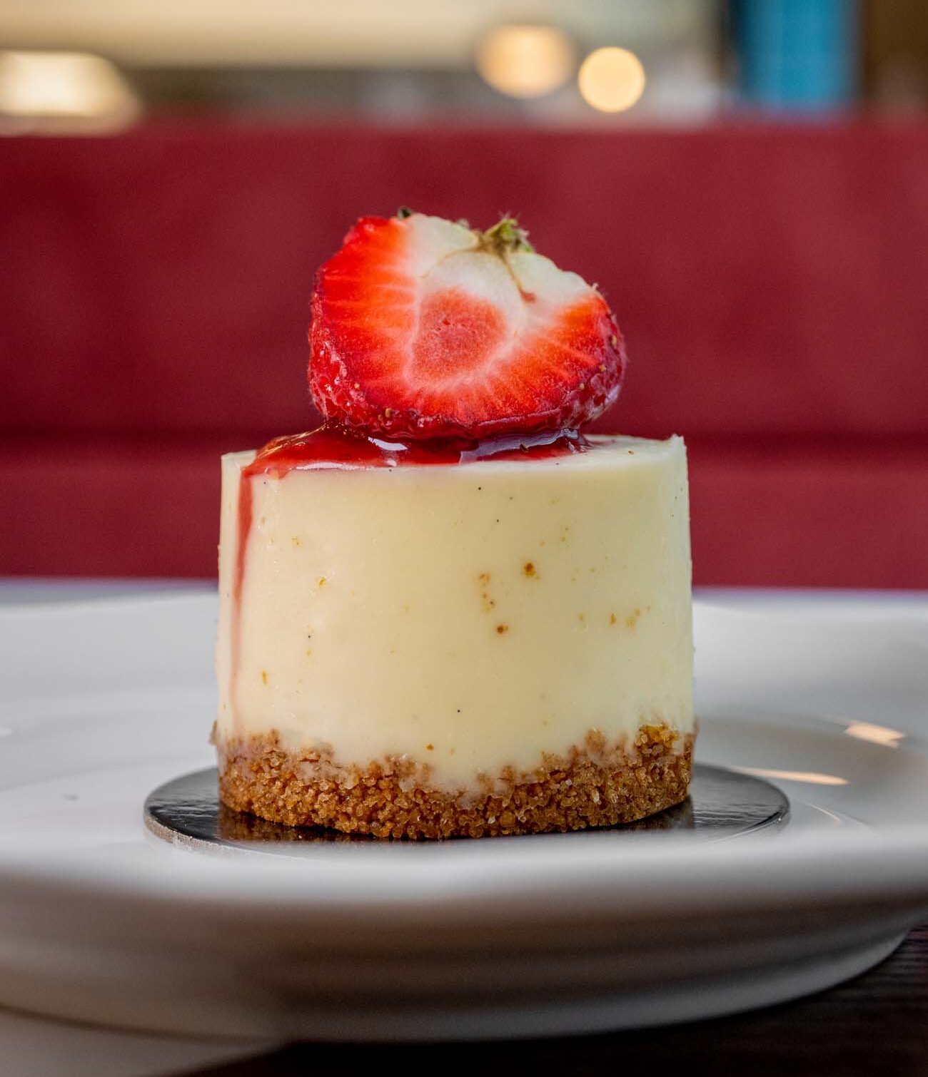 A close-up of an individual-sized vanilla cheesecake with a graham cracker crust, topped with a fresh halved strawberry and sitting on a small black plate against a red background with soft focus.