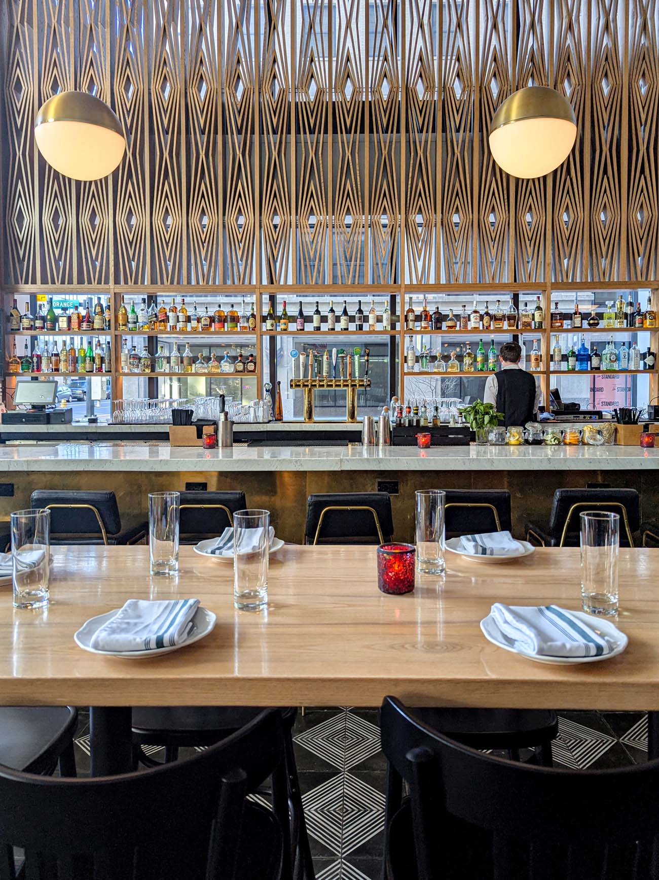 Interior view of Casa Nonna's sophisticated bar featuring geometric diamond-patterned wood wall panels, sleek globe pendant lights, a marble-topped bar counter with black and gold bar stools, and a bartender in black visible behind the well-stocked shelves of spirits.