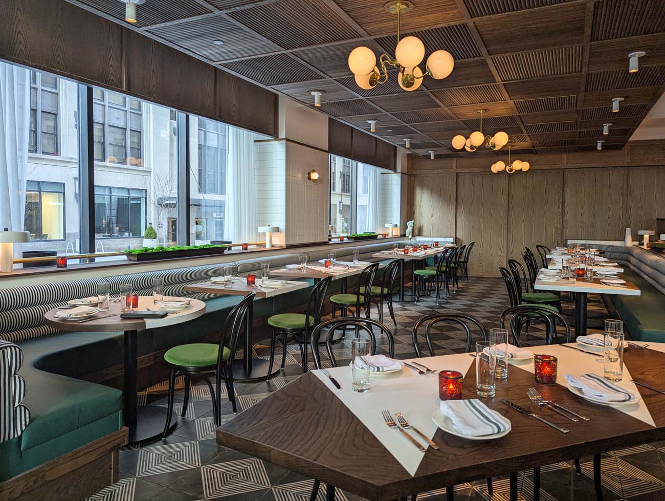 Wide-angle view of Casa Nonna's dining room featuring wood-paneled ceiling with vintage-inspired globe chandeliers, large windows with natural light, black bentwood chairs with green seats, striped banquette seating, and elegant place settings on wooden tables against a geometric tile floor.