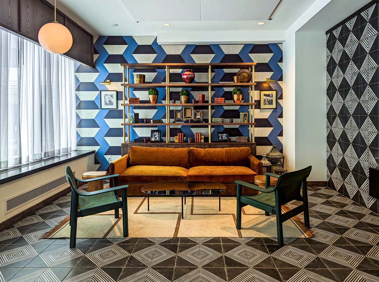 A stylish lounge nook featuring a camel leather sofa, green accent chairs, geometric patterned floor tiles, blue and white geometric wallpaper, and a brass bookshelf filled with decorative objects and books - all creating a sophisticated conversational space.