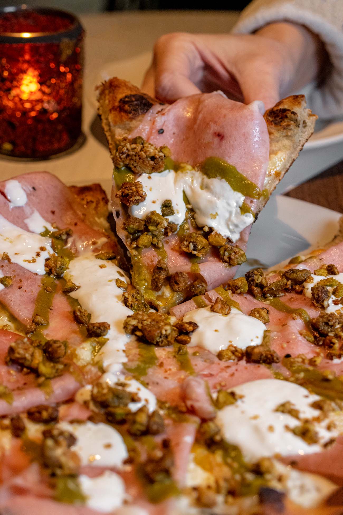 A hand holding a slice of pink mortadella pizza topped with white stracciatella cheese, green herb drizzle, and pistachio crumble, with more pizza visible below showing the same toppings arranged across the entire pie.