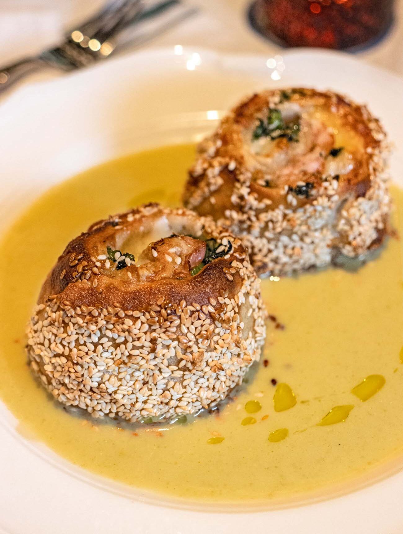 Two sesame-crusted rolls from Casa Nonna sitting in olive oil, featuring twisted layers of porchetta, broccoli rabe and sharp provolone, served with the "utterly addicting long hot cream" sauce mentioned in the article.