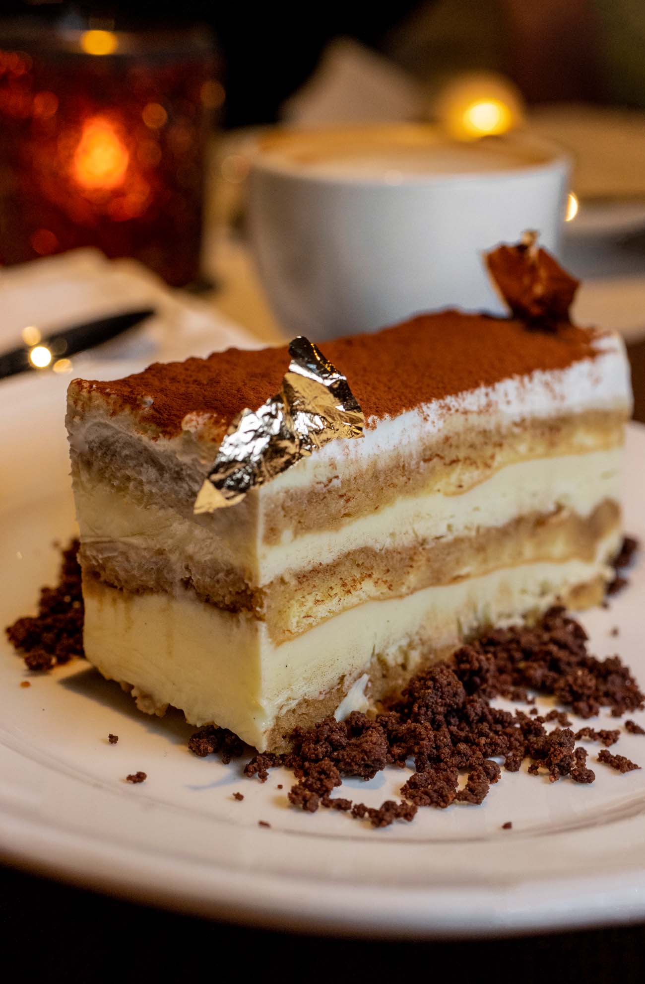 A slice of tiramisu from Casa Nonna on a white plate, showing distinct layers of coffee-soaked ladyfingers, mascarpone cream, and cocoa dusting, garnished with gold leaf as described in the Hotel Du Pont article.