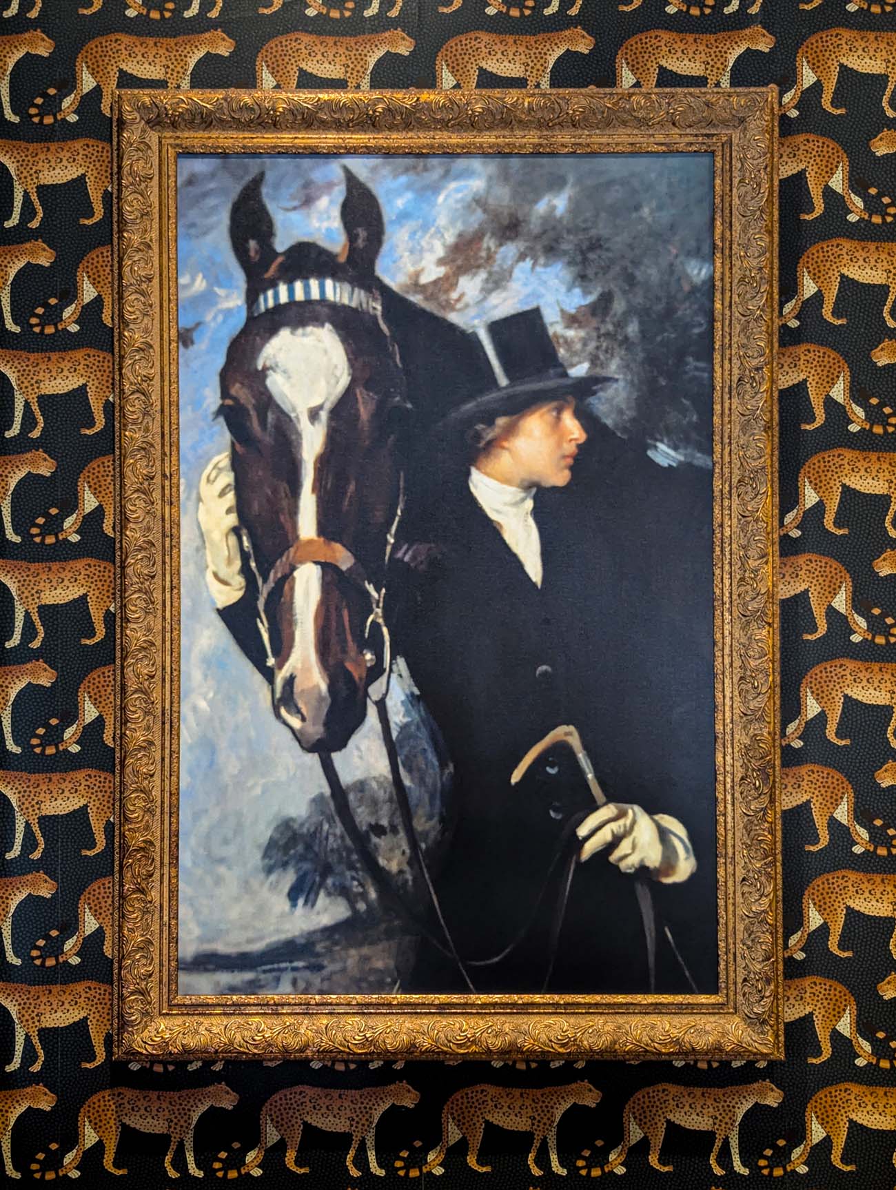 A gold-framed painting of a rider in formal attire with a top hat standing beside a black horse with a white blaze marking, mounted against a leopard-print wallpaper, reflecting the "Horseman" meaning of Le Cavalier and the Brandywine Valley's equestrian 