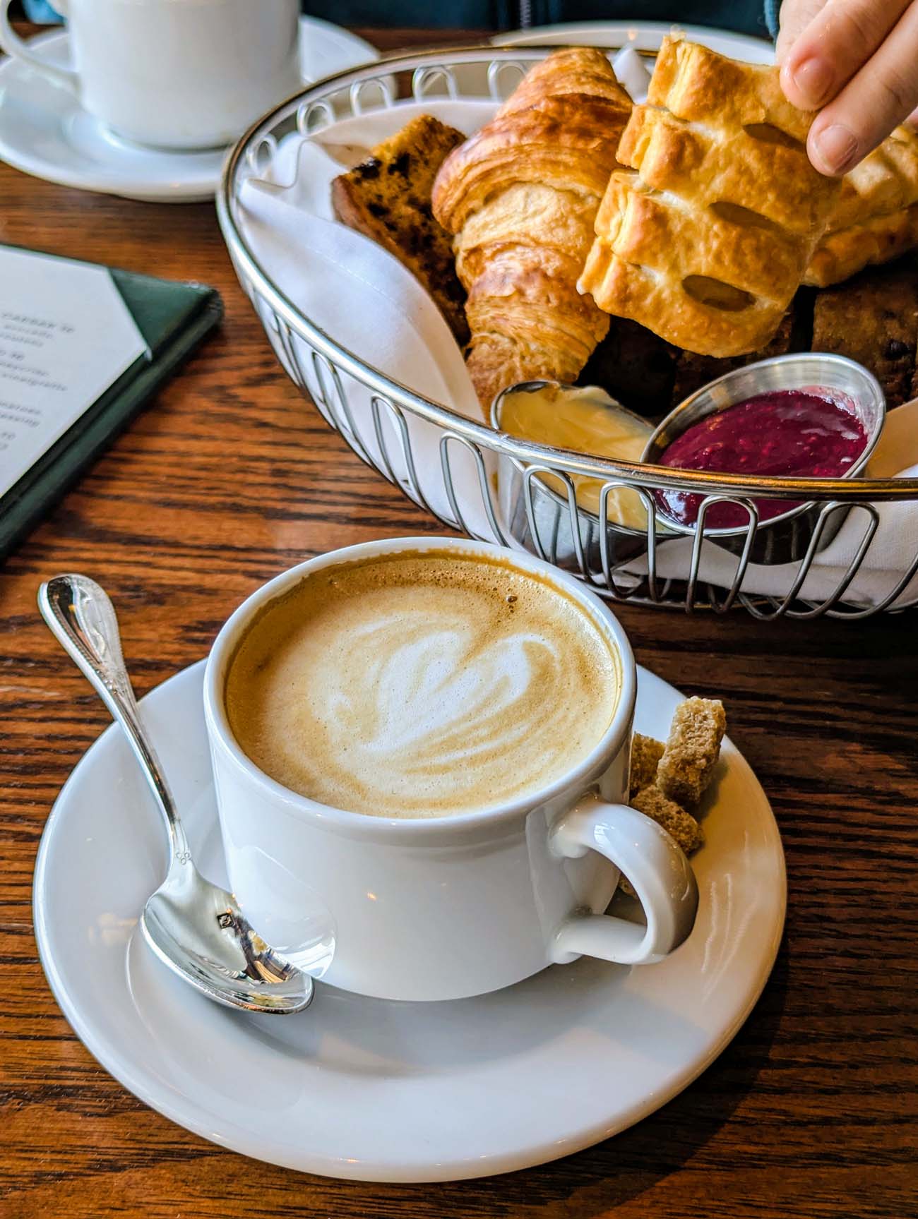 Close-up of a cappuccino with beautiful latte art alongside a silver wire basket containing freshly baked pastries including a golden croissant and other baked goods, with a small dish of house-made jam - the "morning ritual" recommended at Le Cavalier