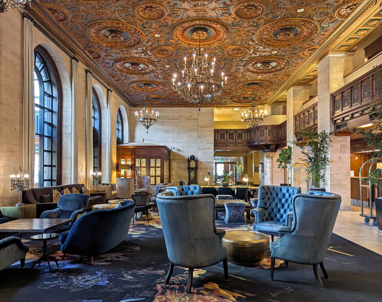  The grand lobby of Hotel Du Pont featuring a spectacular ornate ceiling with gold-detailed medallions, crystal chandeliers, tall arched windows, cream marble walls, and plush blue velvet seating arranged around brass accent tables. The space showcases the hotel's Gilded Age architecture with intricate woodwork and luxurious details dating back to 1913.