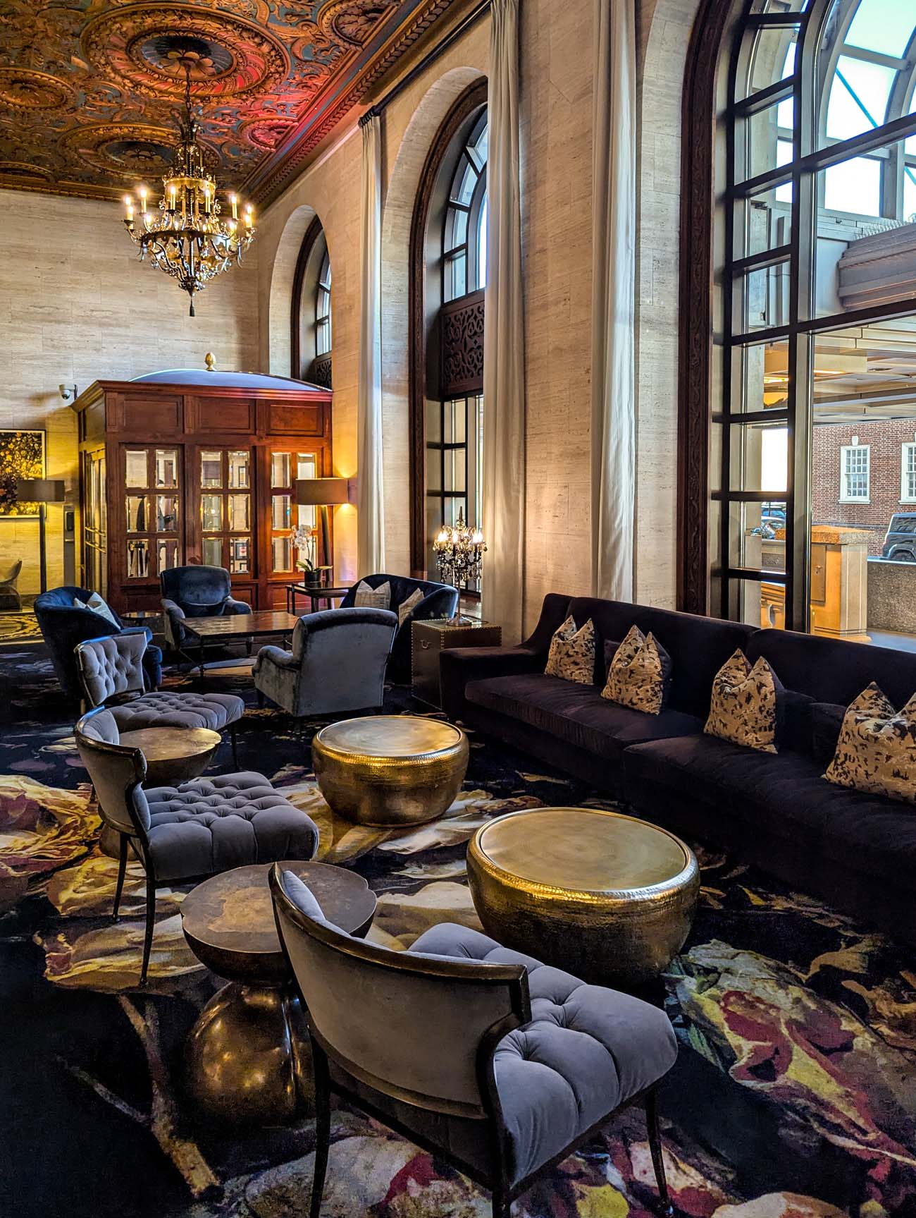 Another view of Hotel Du Pont's magnificent lobby showcasing the ornate painted ceiling, grand crystal chandelier, towering arched windows with drapery, and comfortable seating area with velvet chairs and a dark sofa arranged around brass accent tables. A wooden display cabinet and traditional woodwork highlight the hotel's preserved classic architectural details