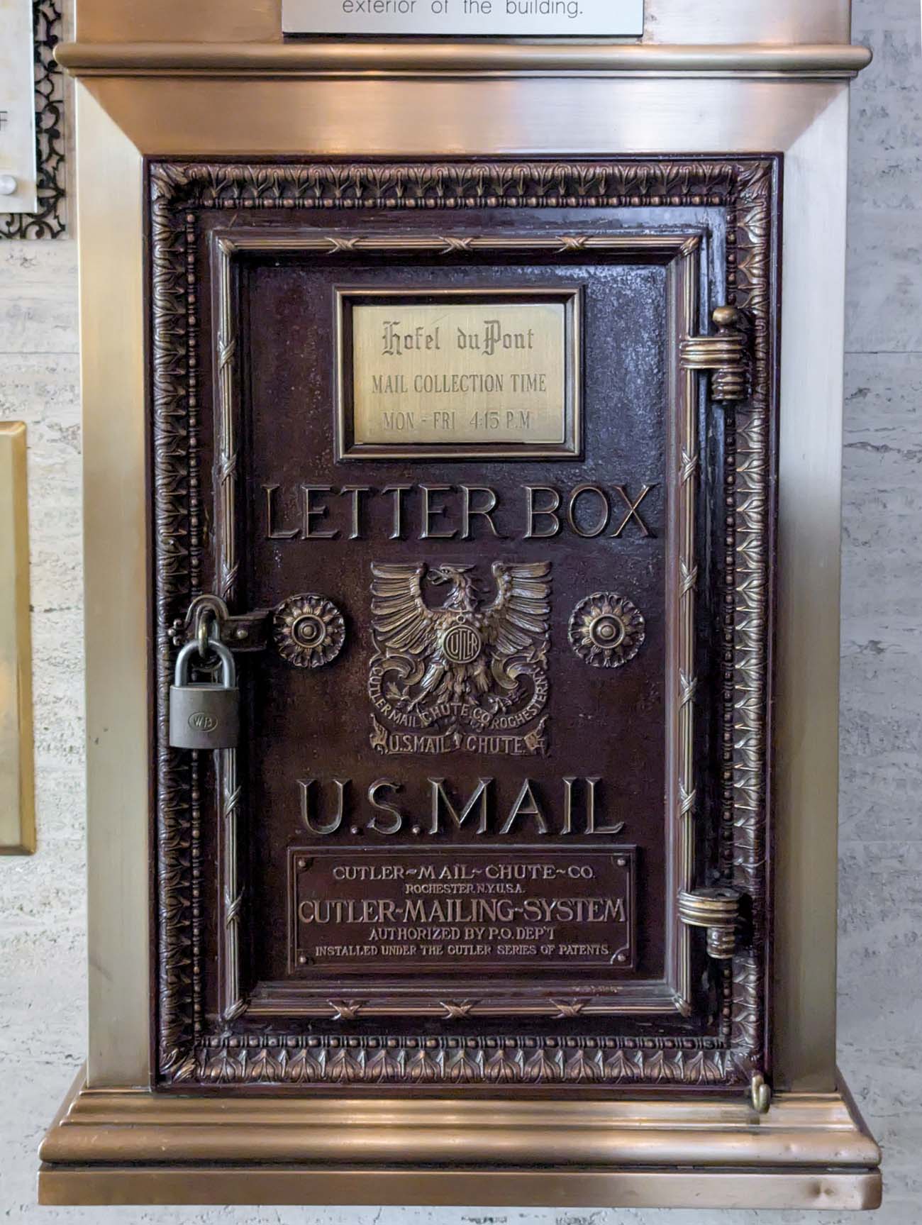 A preserved antique U.S. Mail letterbox installed in Hotel Du Pont's wall, featuring ornate bronze detailing, an eagle emblem, and a gold plaque indicating "Hotel duPont Mail Collection Time Mon-Fri 4:15 P.M." The decorative mail chute, manufactured by Cutler Mail Chute Co., represents the hotel's commitment to preserving unique architectural elements from its 1913 origins.