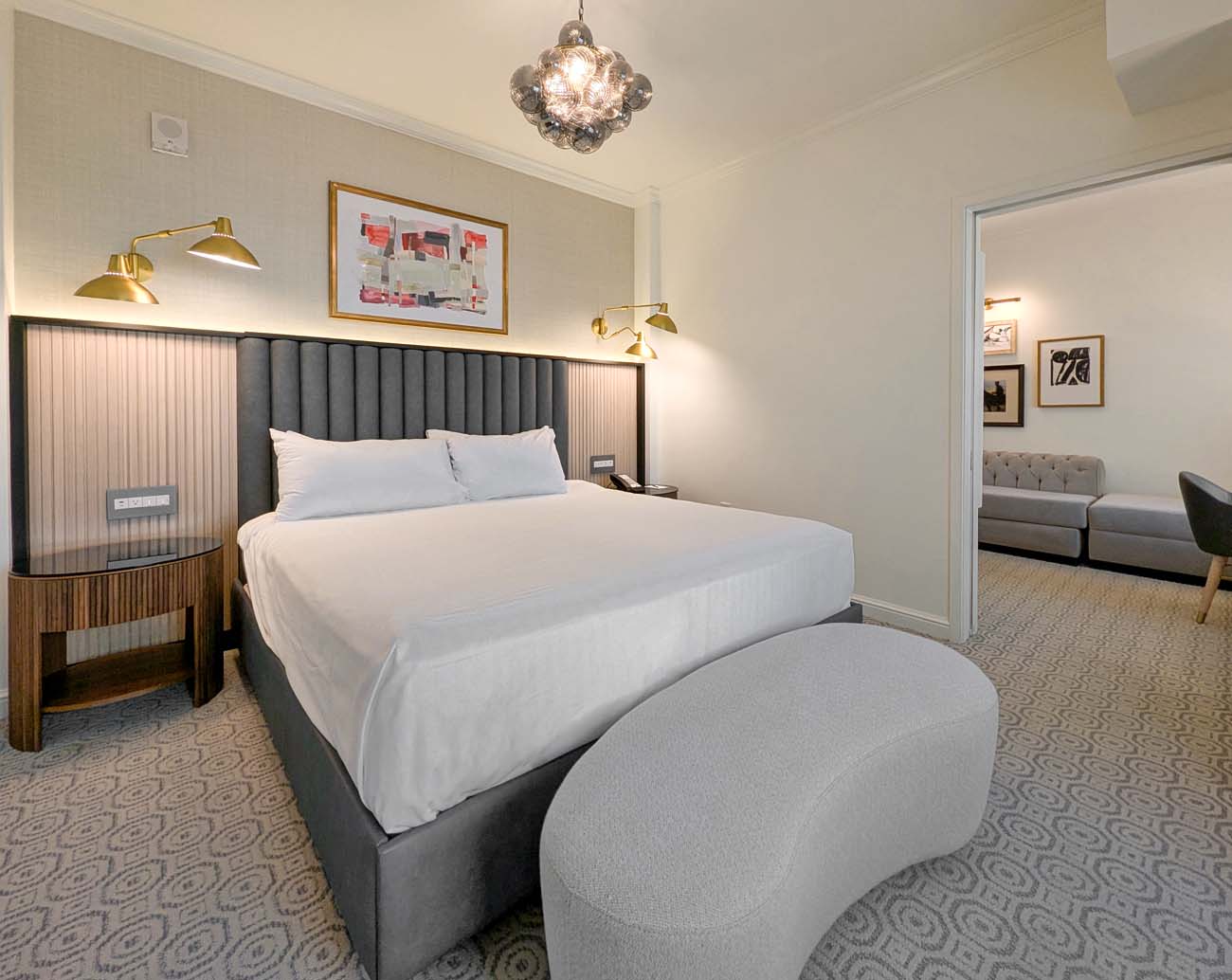 A modernized Hotel Du Pont king bedroom featuring a plush bed with white linens and a gray channel-tufted headboard, contemporary lighting fixtures, abstract artwork, and a view into an adjoining sitting area. The renovated room balances contemporary design with the hotel's historic character through thoughtful details like the crystal pendant light fixture.