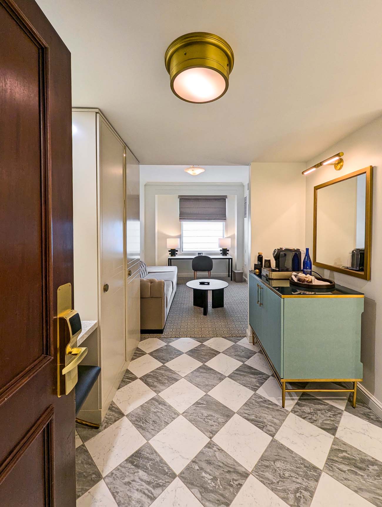 Entryway of a newly renovated Hotel Du Pont suite showcasing a striking checkerboard marble floor, contemporary mint-green cabinet with gold accents, and view into a sitting area with beige sofa and modern furnishings. The spacious accommodation features elegant architectural moldings and brass fixtures that complement the hotel's historic character.