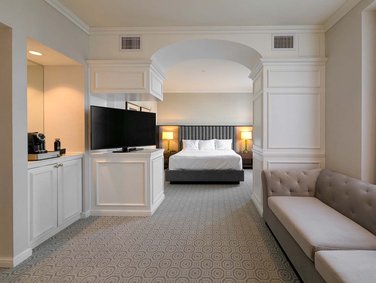 A view through an archway in a Hotel Du Pont suite, looking from a living area with light gray sofa toward the bedroom with king-sized bed, gray channel-tufted headboard, and bedside lamps. The renovated suite features classic white paneling, carpeted floors with geometric patterns, and a flat-screen TV, blending modern amenities with the hotel's historic charm