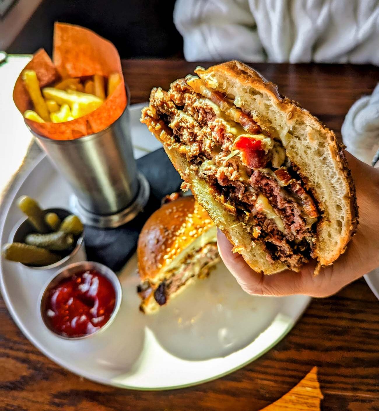 A hand holding half of Le Cavalier's famous Le Cheeseburger, revealing two juicy prime beef patties with bacon and sauce on a buttery toasted brioche bun. The plate below contains the other half of the burger served with golden shoestring fries in a metal cup, pickle spears, and a side of ketchup - showcasing the upscale comfort food available at the hotel.