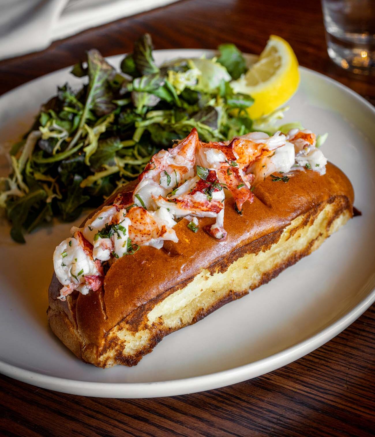 A golden toasted roll overflowing with chunks of fresh lobster meat garnished with herbs, served on a white plate alongside a bright green salad and lemon wedge. This elegant preparation demonstrates Le Cavalier's approach to classic dishes, offering guests both hot buttered and cold Duke's Mayo preparations as mentioned in the article.