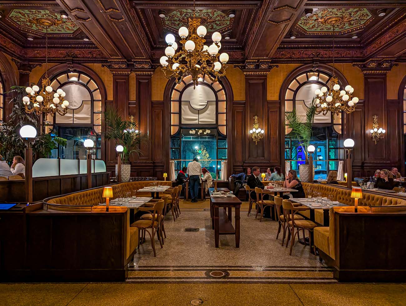 The stunning interior of Le Cavalier restaurant (formerly the Green Room) at Hotel Du Pont, showcasing the breathtaking ornate ceiling with painted medallions, original crystal chandeliers, grand arched windows, rich wood paneling, and gold velvet banquettes. Diners enjoy meals at tables set with white tablecloths in this historically significant dining space that blends old-world charm with contemporary elements.