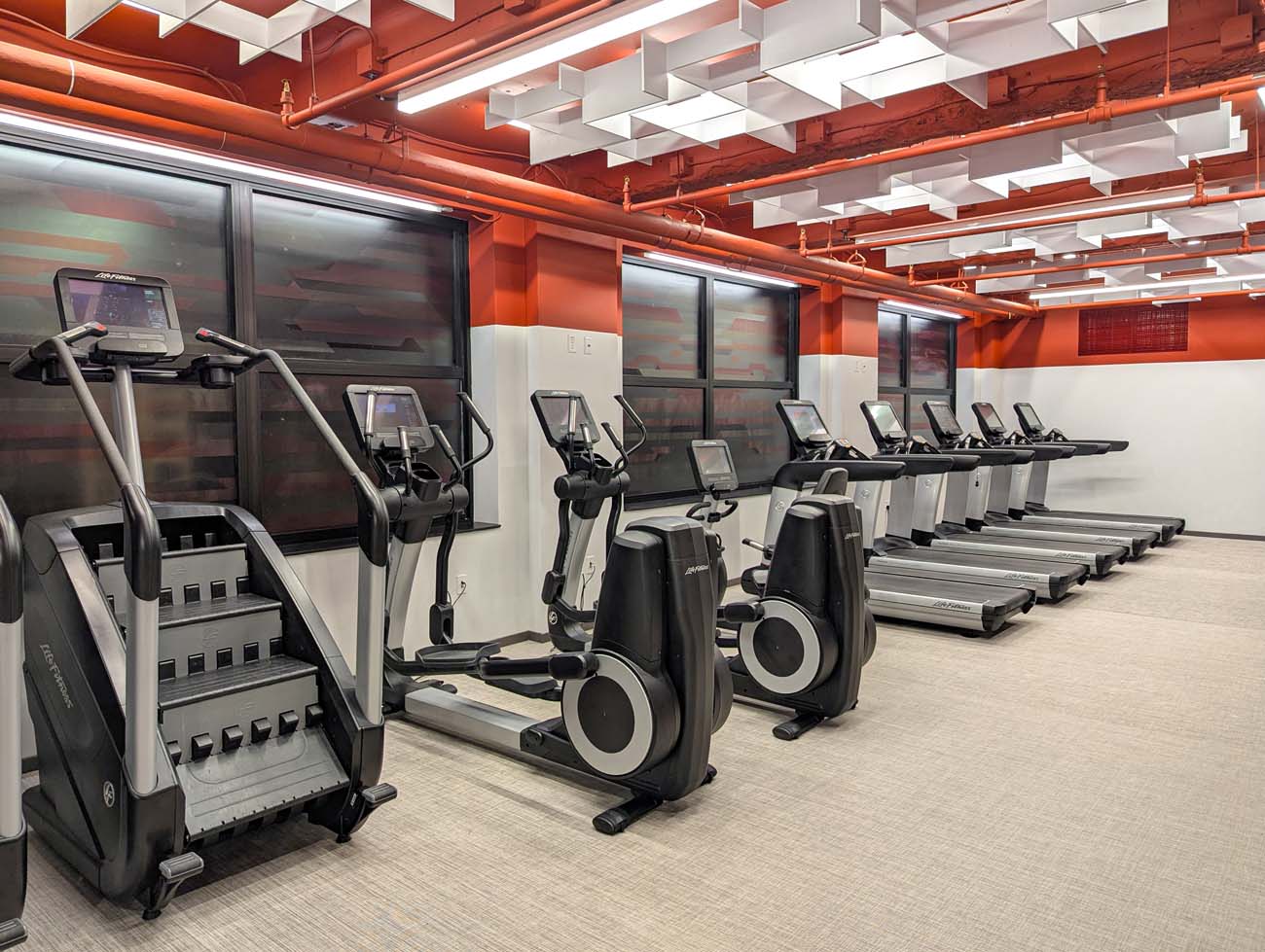 The modern 24-hour fitness center at Hotel Du Pont featuring a row of treadmills, ellipticals, stair climber, and Peloton bikes against frosted windows with orange accent walls and white acoustic ceiling panels. The well-equipped gym, located on the 2nd floor, provides hotel guests with comprehensive workout options including cardio machines and strength training equipment.