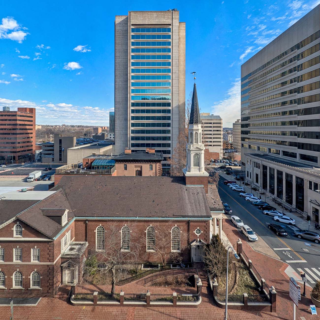 Aerial view of downtown Wilmington with Hotel Du Pont (the tall concrete building in center) dominating the skyline against a bright blue sky. A historic brick church with white steeple stands in the foreground, illustrating the hotel's prime location in Wilmington's business district as described in the article.