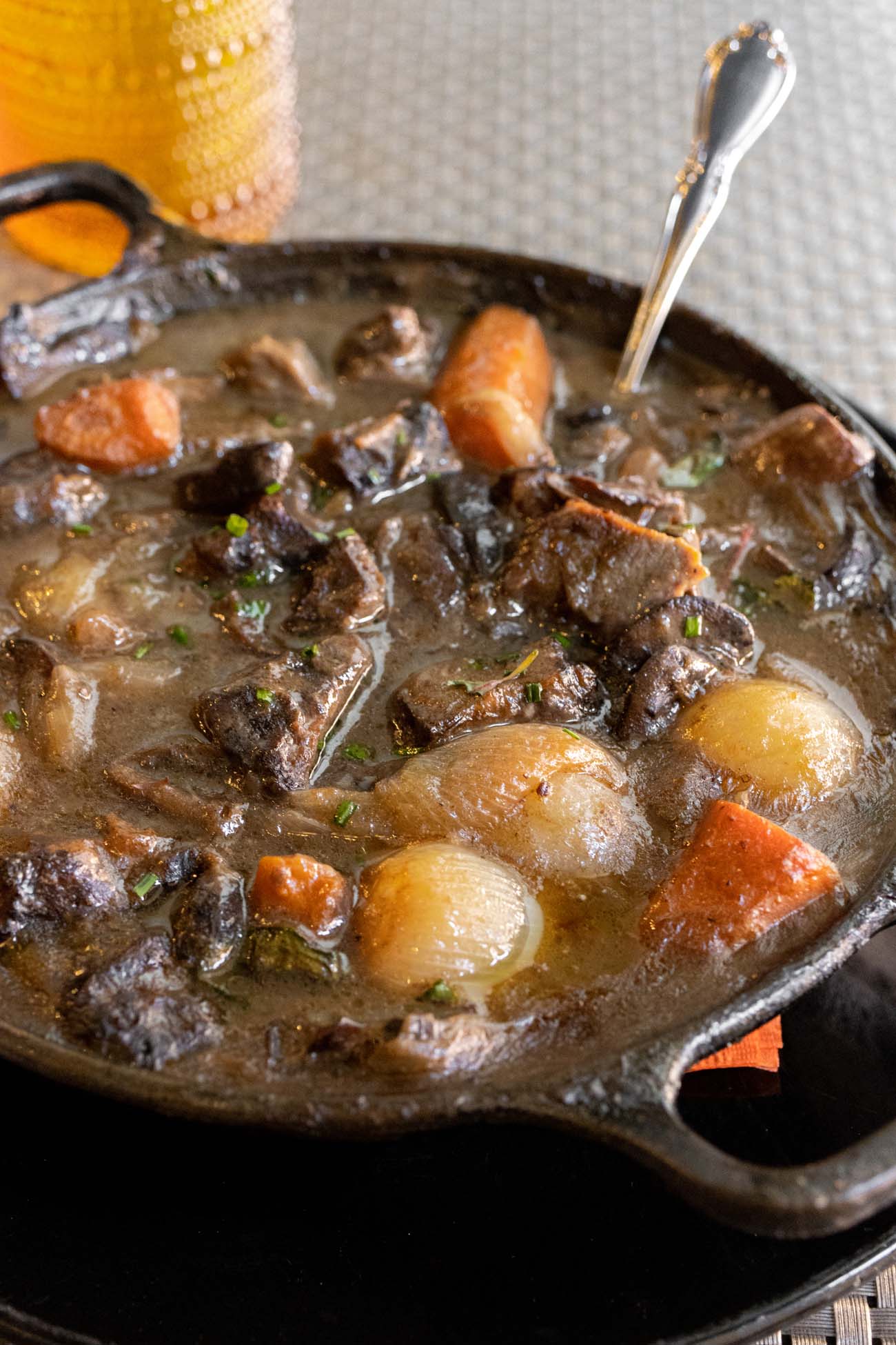 A close-up photograph of traditional French Boeuf Bourguignon served in a rustic cast iron skillet. The rich, dark brown stew features tender chunks of beef, pearl onions, and carrots in a silky wine sauce. Fresh herbs are sprinkled on top, and a silver spoon rests in the dish. The stew is steaming hot and presented on a dark surface with a glimpse of a yellow beverage in the background.