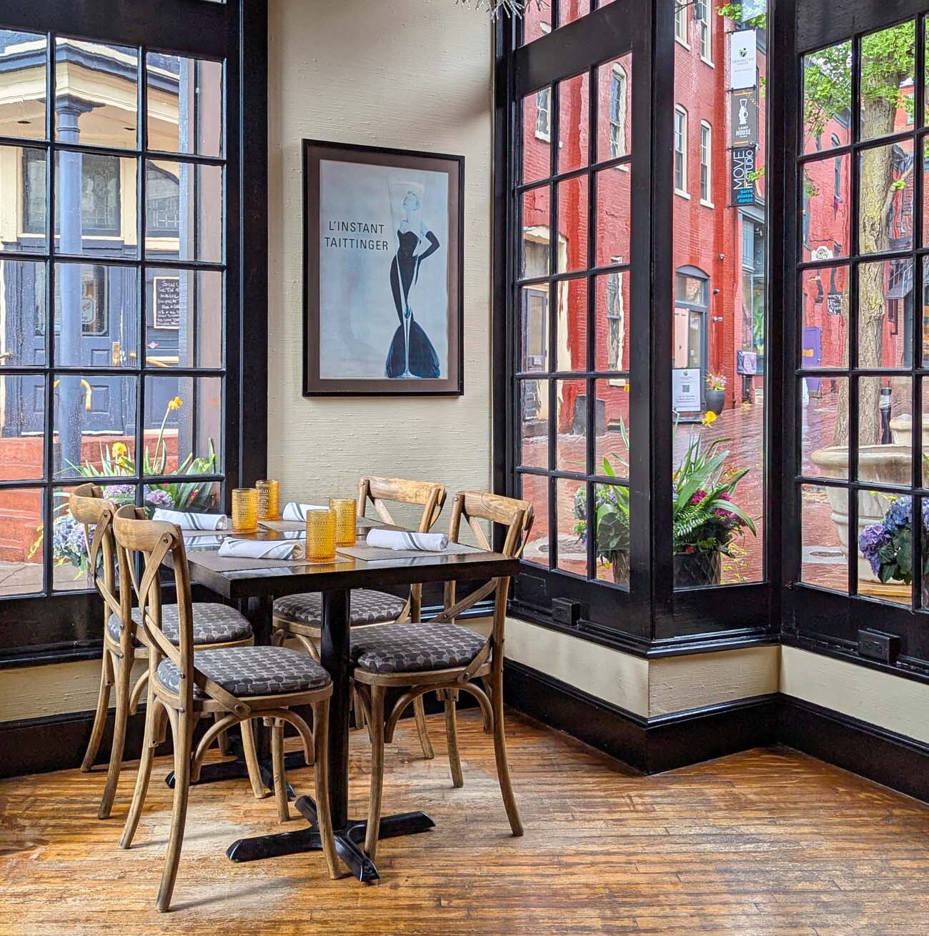 A bright, airy corner table at C'est La Vie French bistro in downtown Lancaster. The space features floor-to-ceiling black-framed windows on two sides, offering views of historic red brick buildings across the street. A Taittinger champagne poster decorates the cream-colored wall. The wooden table is set with amber-colored glasses and white napkins, surrounded by bistro chairs with patterned cushions. Hanging wire light fixtures and potted plants add to the French café atmosphere. Sunlight streams through the windows, illuminating the worn wooden flooring.