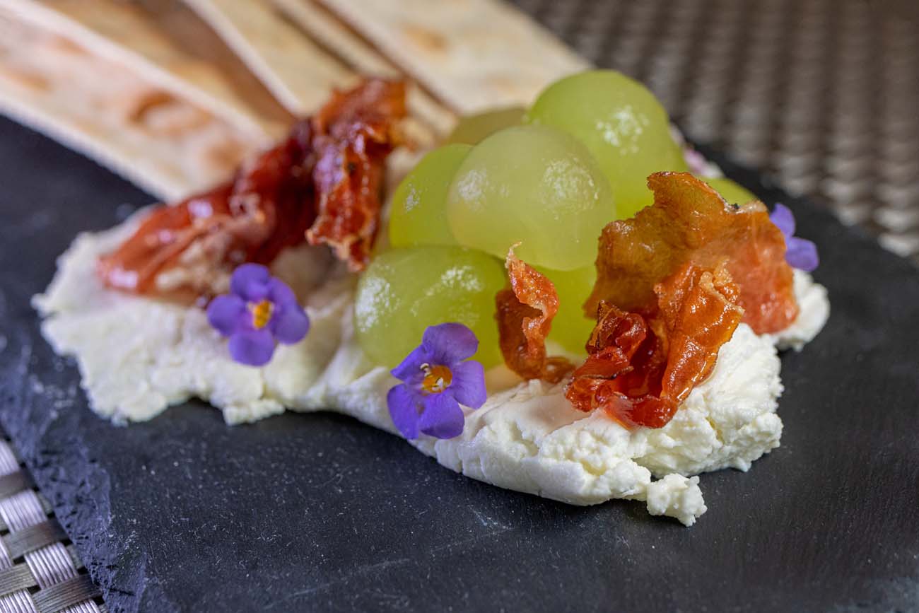 A beautifully composed appetizer from C'est La Vie French bistro in Lancaster, showcasing their artistic presentation and French culinary techniques. A creamy white spread of goat cheese forms the base on a slate plate, topped with four glistening melon balls, thin crispy pieces of prosciutto, and delicate purple edible flowers. A thin flatbread or lavash is partially visible at the left edge of the frame. The dish exemplifies C'est La Vie's attention to detail and balance of flavors, combining sweet, salty, and creamy elements in an elegant presentation that reflects the restaurant's authentic French bistro cuisine.