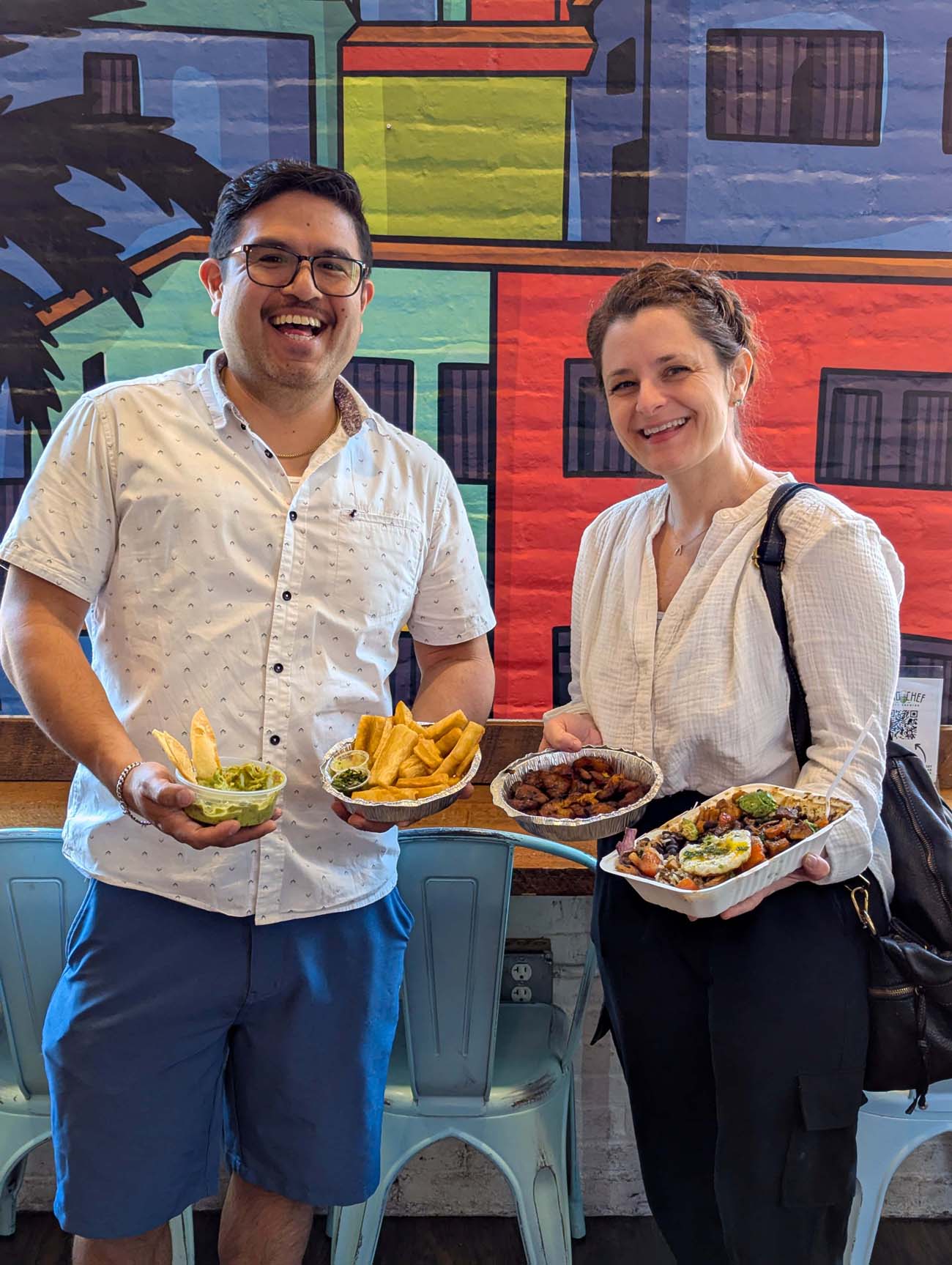 Two people smiling and holding food from Chellas Arepa Kitchen in Lancaster. On the left, a man in a white patterned short-sleeve shirt (Luis, the owner) holds a container of guacamole and a plate of plantain chips. On the right, Heather from Better Living in a white top holds two aluminum containers with various Venezuelan dishes. They're standing in front of a colorful mural with bright blue, red, yellow, and green geometric designs that reflect South American artistic influences. The casual, welcoming atmosphere showcases the authentic cultural experience offered at this hidden gem on North Queen Street.