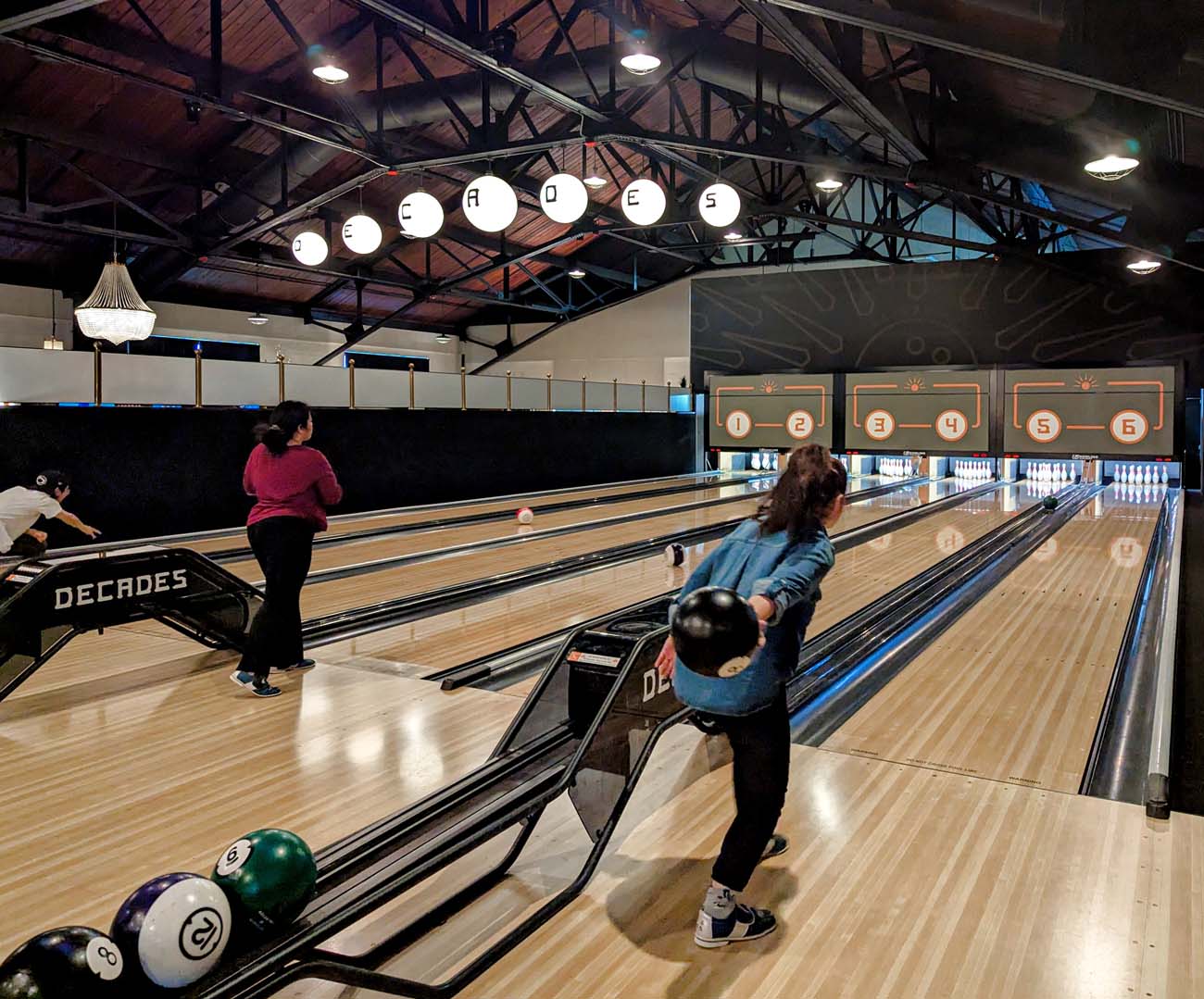 Interior view of the boutique bowling alley at Decades in downtown Lancaster. The image shows several polished wooden bowling lanes with two bowlers in action. One woman in a pink shirt is walking toward the lane to bowl, while another in a blue denim jacket is poised to release her ball. Overhead, the name "DECADES" is illuminated in circular white signs hanging from the rustic wooden rafters of the renovated 95-year-old Stahr Armory building. The stylish retro-modern atmosphere features vintage-inspired lane markers and a chandelier visible in the distance. The boutique setup offers a more intimate bowling experience than traditional alleys, aligning with Decades' concept of blending nostalgia with upscale food and drink offerings.