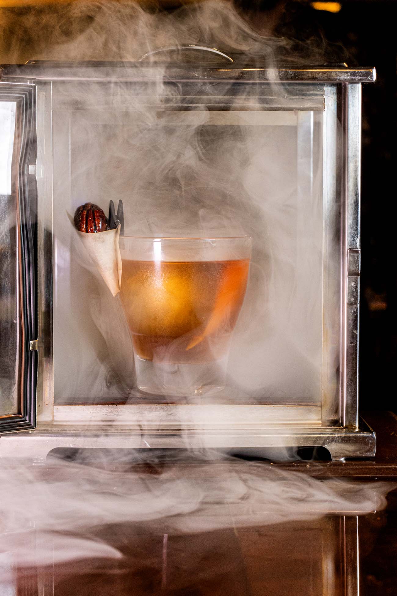 A theatrical cocktail presentation at Decades showing their Toasted Pecan Smoked Old Fashioned inside a glass smoking chamber. The amber-colored bourbon cocktail sits in a lowball glass within a transparent box filled with swirling white smoke. A decorative garnish of what appears to be a caramelized pecan or spice is visible on the side of the glass. The smoke adds both flavor and dramatic effect to the drink, demonstrating the innovative craft cocktail program that complements Decades' retro gaming experience. This particular cocktail features Elijah Craig bourbon, toasted pecan syrup, and angostura bitters, smoked tableside for an immersive sensory experience.