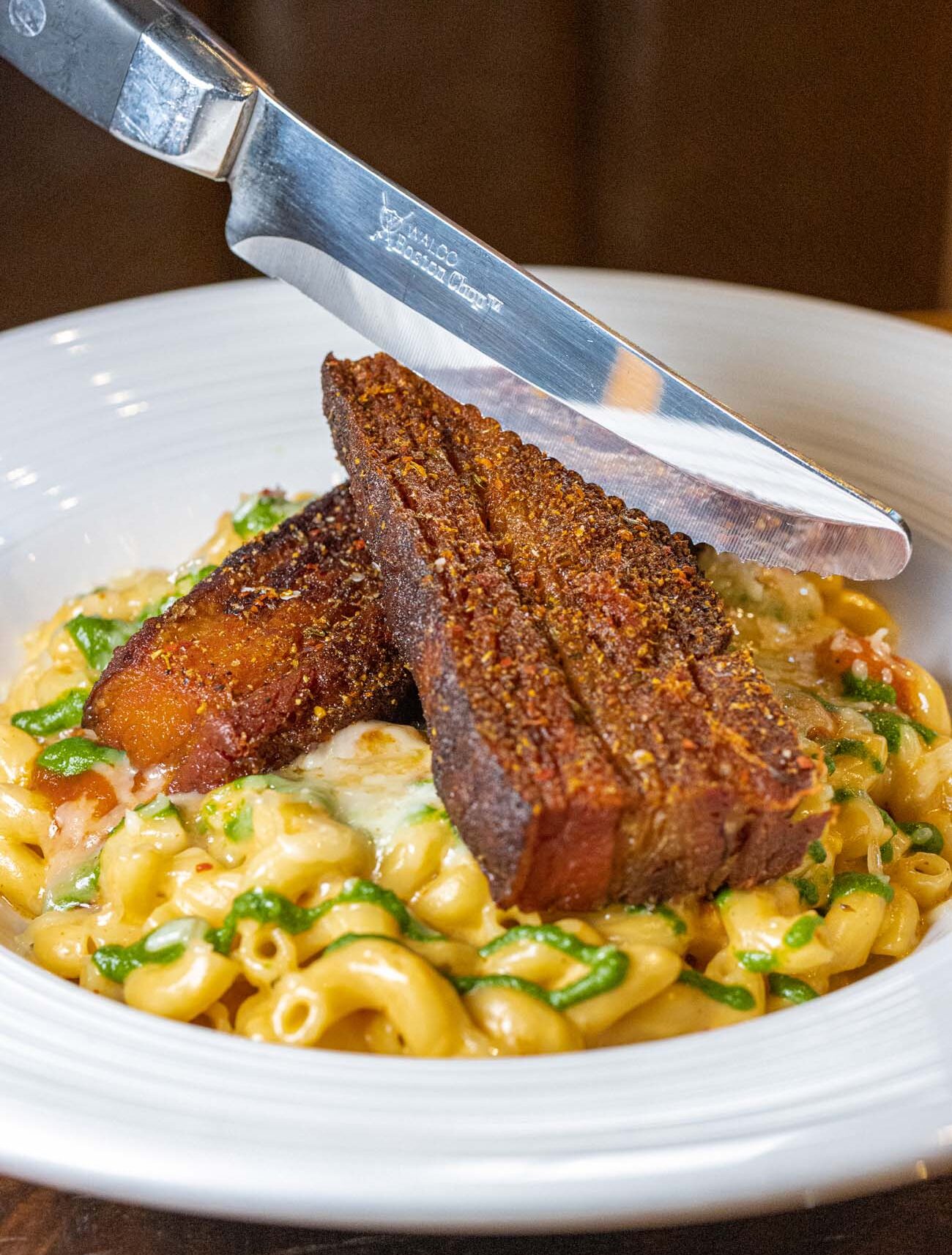 Close-up of an elegantly plated dish at Double C restaurant in Lancaster, featuring two triangular pieces of seared pork belly with a deeply caramelized, spice-crusted exterior being cut with a steak knife. The crispy pork belly rests atop a bed of creamy mac and cheese pasta with fresh green herbs incorporated throughout. The dish is served in a white bowl against a dark background. This presentation exemplifies Double C's "Keystone Casual" approach—ethically sourced, pasture-raised meats prepared with culinary precision but served in a relaxed setting. The knife bears the restaurant's or manufacturer's engraving, highlighting the attention to quality details. This dish appears to be their famous Double C Bowl with crispy pork belly, which combines spicy mac and cheese, fire-roasted tomato sauce, chimichurri, and shredded cheese.