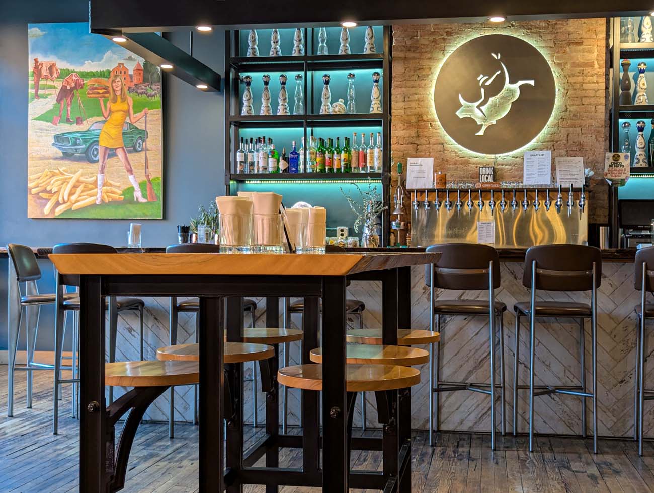 Interior view of Double C restaurant and bar in downtown Lancaster. The stylish space features a live-edge wooden bar table with modern bar stools in the foreground. Behind the bar, turquoise-lit shelves display an impressive collection of spirits and craft liquors. The exposed brick wall on the right showcases an illuminated circular logo that appears to be the restaurant's emblem, while the blue wall on the left displays a vibrant mural depicting a retro scene with a woman in a yellow dress standing next to a vintage car and french fries. This playful artwork reflects Double C's "Keystone Casual" concept mentioned in the blog post, where seriously good, responsibly sourced food is served in a relaxed, no-attitude space. The sophisticated blend of historical architecture with contemporary design elements embodies chefs Sean Cavanaugh and Michael Carson's approach to ethical sourcing with creative presentation.
