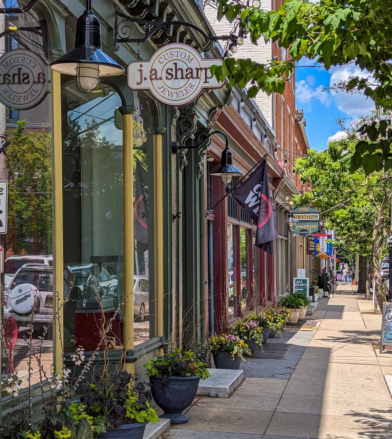 A sunlit view down North Queen Street in downtown Lancaster showing well-preserved historic storefronts with their original architectural details. In the foreground is J.A. Sharp Custom Jeweler with its distinctive hanging sign, housed in a sage green painted storefront with ornate Victorian-era details. The sidewalk is lined with decorative planters filled with colorful flowers and shrubs. Looking down the street, other boutique shops with colorful awnings and signs are visible, along with leafy trees providing shade. The scene showcases Lancaster's charming blend of historic preservation and small business vitality, highlighting the 300 Block area mentioned in the travel guide as a prime shopping destination. The bright blue sky and sunshine create an inviting atmosphere for exploring these independent retailers.