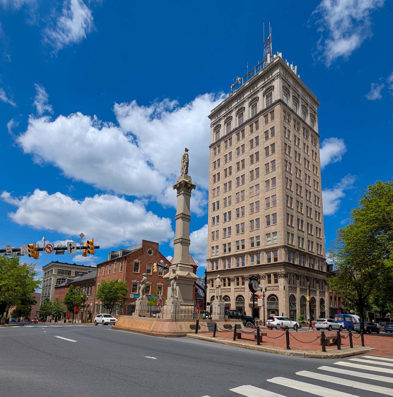 A street-level view of downtown Lancaster's Penn Square, dominated by the historic Griest Building—a 14-story beige limestone skyscraper that was once the tallest building between Philadelphia and Pittsburgh. In the foreground is a crosswalk leading into the square, where a tall stone monument or soldiers' memorial stands. The classical architecture of the Griest Building contrasts with the smaller red brick buildings surrounding the square. Traffic lights and street signs are visible at the intersection, and the scene is captured on a beautiful day with vibrant blue skies and fluffy white clouds. This view showcases the heart of downtown Lancaster where many historic events have taken place, including serving as the U.S. capital for one day during the Revolutionary War. The image highlights the walkable nature of downtown Lancaster with its grid-pattern streets that have remained largely unchanged since the city's founding.