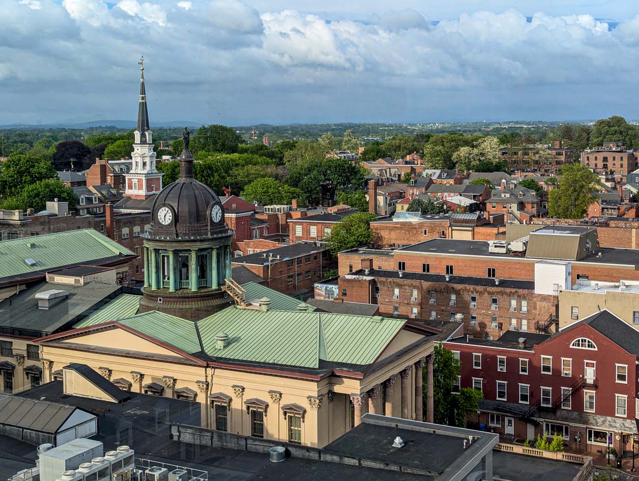 A sweeping aerial view of downtown Lancaster's historic skyline with the distinctive domed clock tower of the old courthouse or city hall in the foreground. The dome features a copper-green patina with a clock face visible on its side. Beyond the dome, the scene captures Lancaster's characteristic red brick buildings spanning several centuries of architecture, punctuated by church steeples and varied rooflines. In the distance, rolling hills and the surrounding countryside are visible under a dramatic sky with fluffy clouds. This perspective perfectly illustrates the travel guide's description of Lancaster's seamless transition from vibrant urban center to peaceful rural landscape. The image shows the density of historic structures within the city's compact seven square miles, highlighting why Lancaster's National Register Historic District is one of the largest in the United States with over 14,000 buildings.