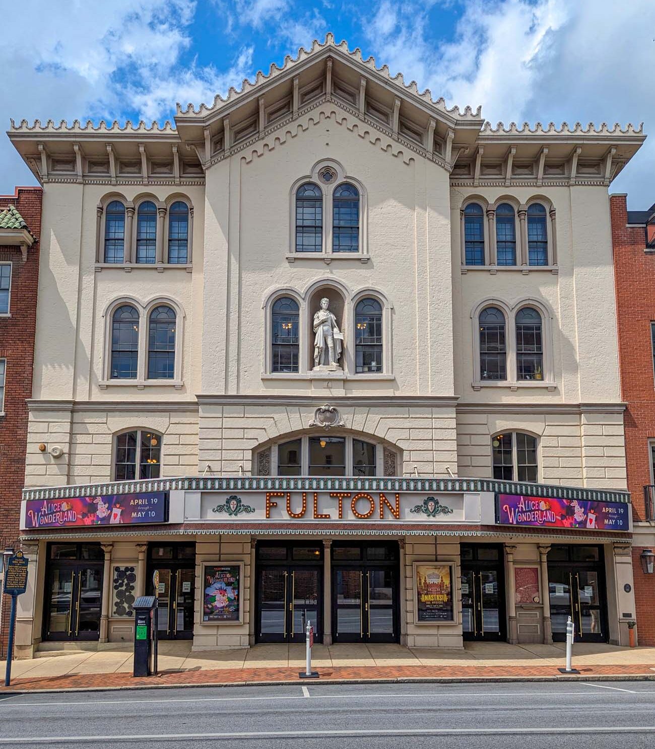 ront exterior view of the historic Fulton Theatre in downtown Lancaster, showcasing its distinctive Victorian architecture. The cream-colored building features ornate decorative elements, arched windows, and a prominent central statue niche containing a classical figure. The theater's marquee displays "FULTON" in illuminated letters along with posters advertising "Alice in Wonderland" running from April 19 to May 10. Built in 1852 on the foundation of Lancaster's pre-Revolutionary jail, this National Historic Landmark exemplifies the preservation of Lancaster's rich cultural heritage. The elaborate architectural details, including decorative cornices and window treatments, reflect the building's importance as a cultural landmark that has hosted performers from Mark Twain to modern Broadway productions. The pristine condition of the facade demonstrates the results of the recent $19 million renovation mentioned in the travel guide.