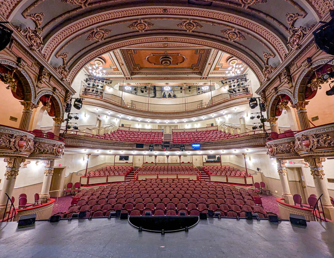 A stunning view of the Fulton Theatre's auditorium interior as seen from the stage looking toward the audience seating. The theater features elaborate Victorian ornamentation with ornate gold-detailed arches, decorative plasterwork on the ceiling, and rich red velvet seating throughout the orchestra and balcony levels. The curved balcony wraps around the auditorium, creating an intimate performance environment despite the theater's size. Modern lighting and sound equipment has been seamlessly integrated into the historic structure, demonstrating the $19 million renovation mentioned in the travel guide. The empty red seats await theatergoers, with the stage visible in the foreground. This view showcases why the Fulton Theatre is considered a crown jewel of Lancaster's cultural scene and highlights the blend of historical significance (as America's oldest continuously operating theater) with modern production capabilities.
